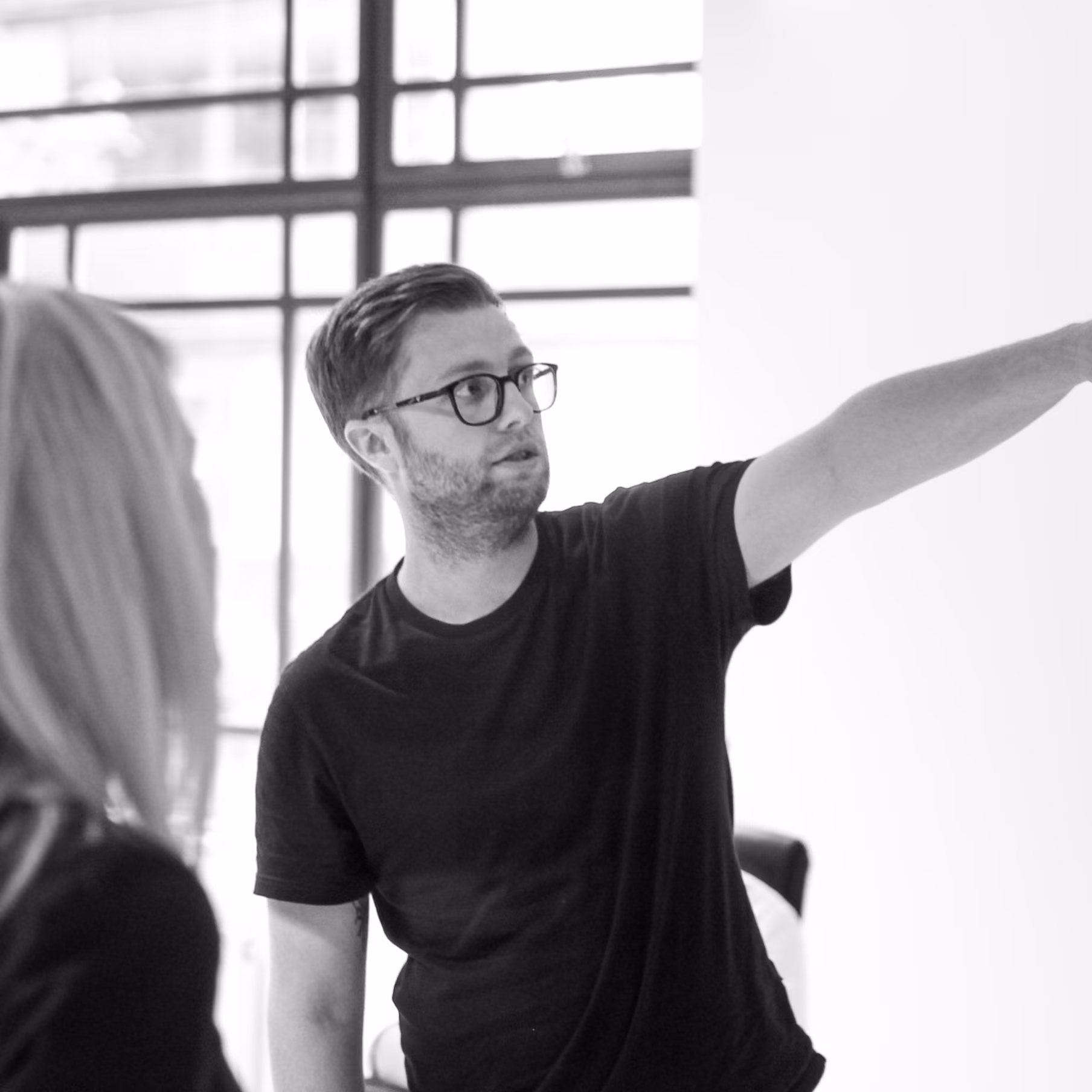 A man wearing glasses and a black t-shirt points to something off to the right while speaking to a woman with blonde hair in a bright room with large windows.