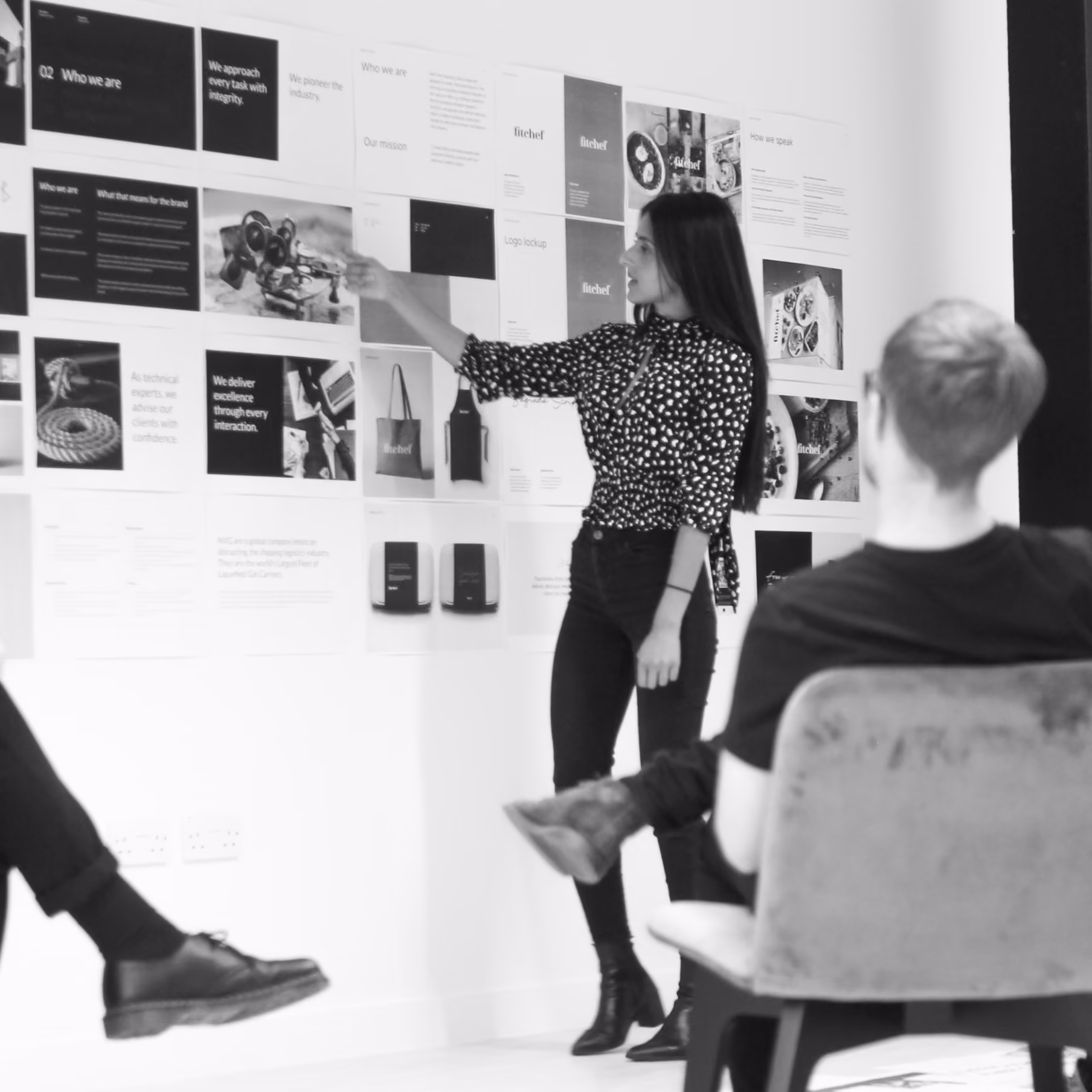 A woman in a polka dot blouse stands and points at various documents and images pinned to a white wall during a meeting, while two seated people listen attentively.