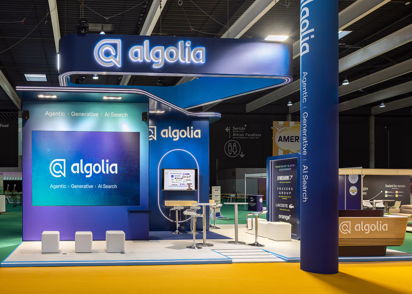 Modern Algolia booth with blue branding, digital screens, and white stools at a trade show.
