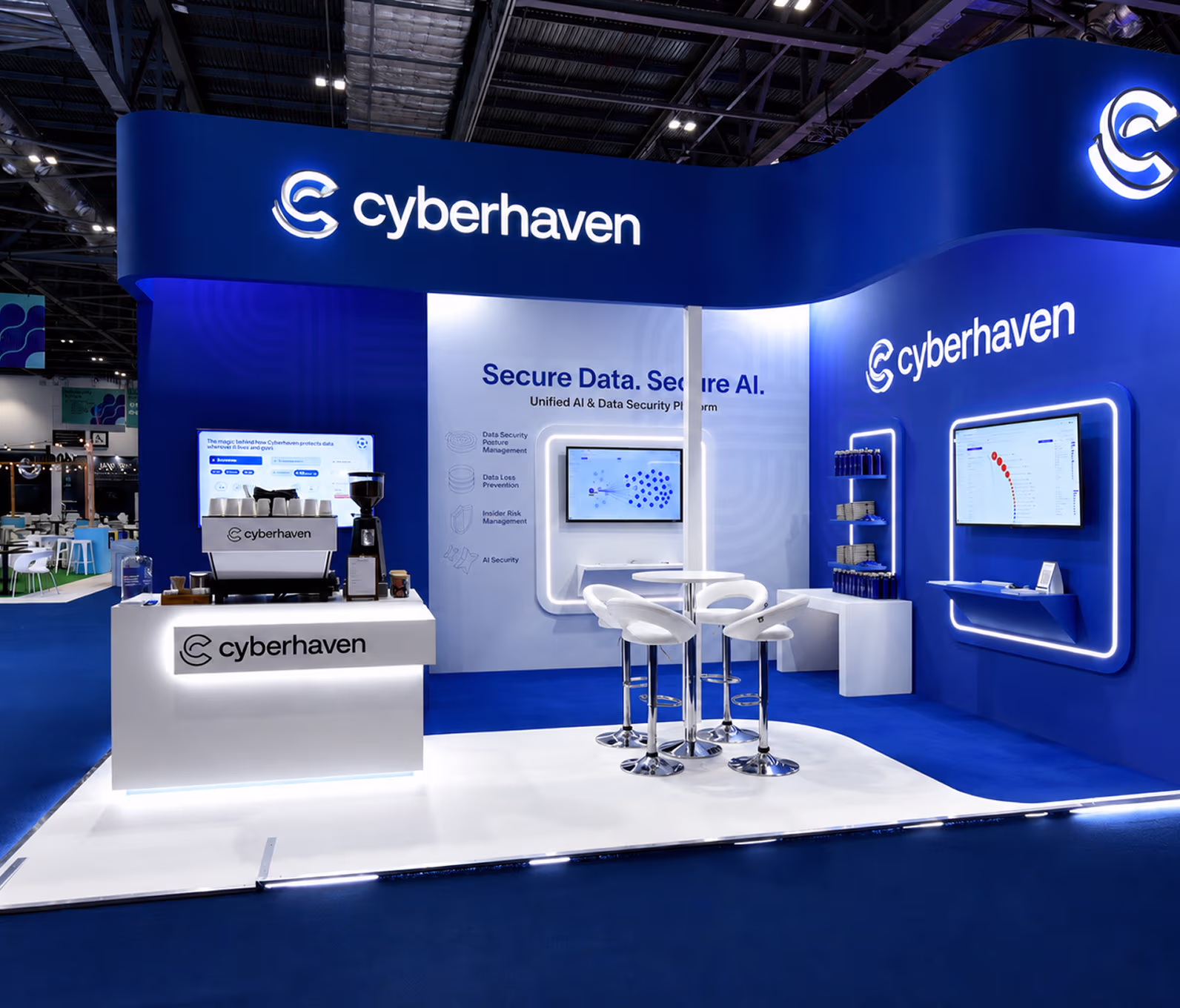 Cyberhaven exhibit booth at conference with blue and white branding, featuring logos, screens displaying data and charts, a coffee machine counter, and a seating area with white bar stools.