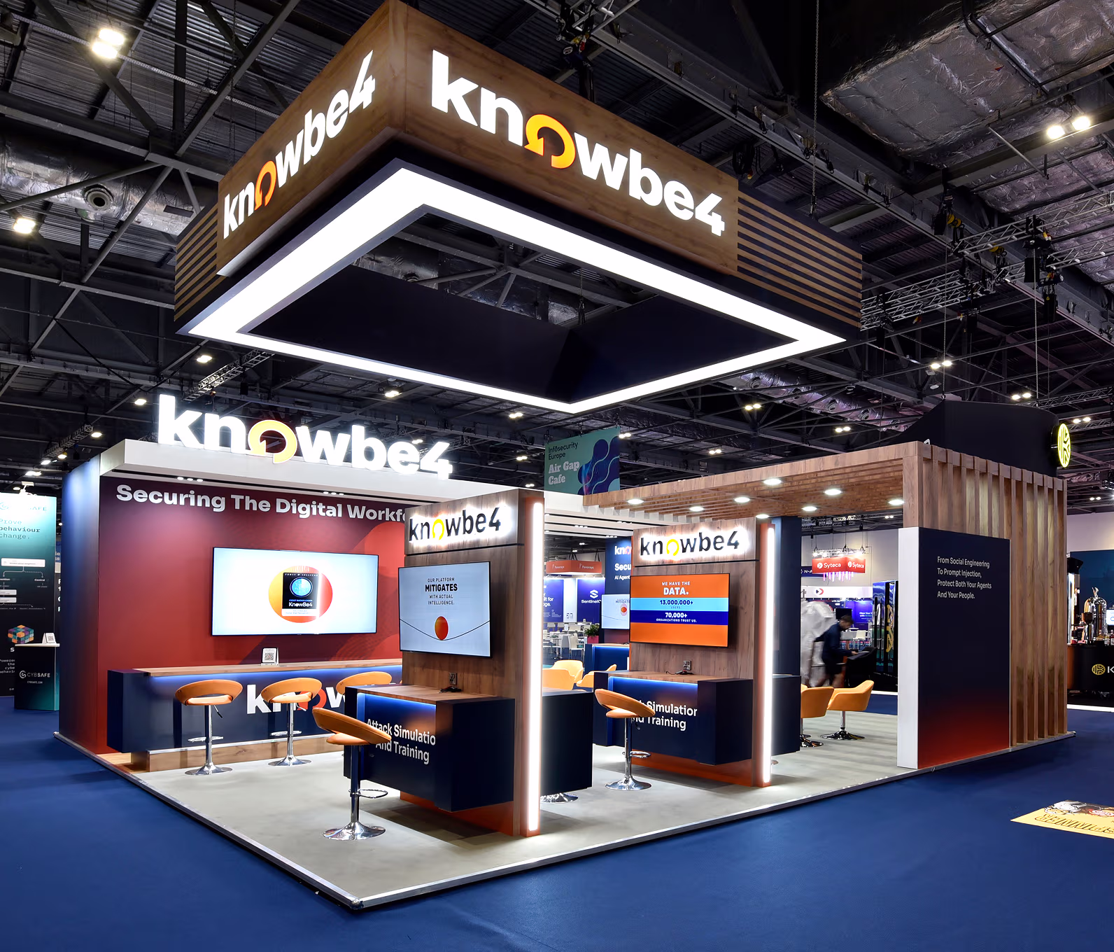 KnowBe4 trade show booth with illuminated branding, multiple screens, orange stools, and seating areas on blue carpet in an exhibition hall.