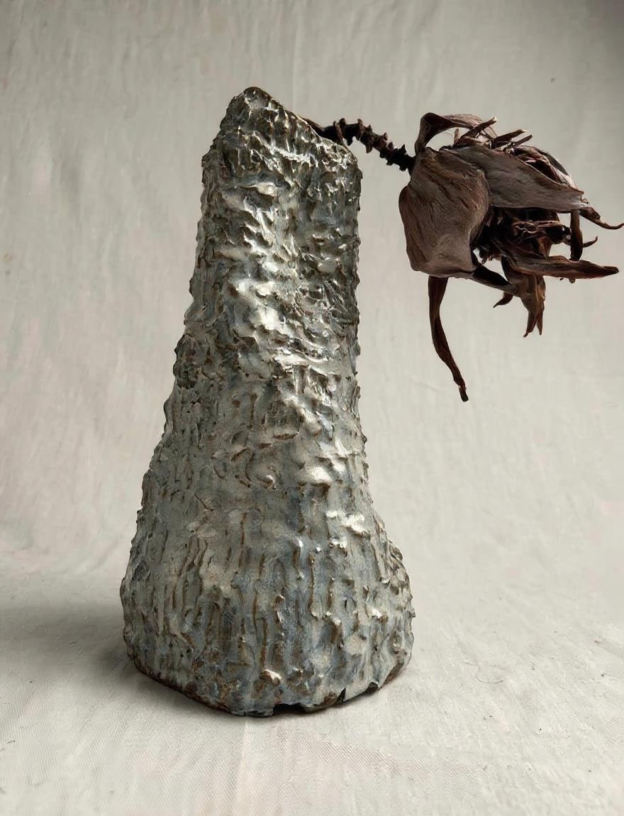 Tall tapering hand-built ceramic vessel with heavy vertical texture in grey and green tones, holding a dried botanical stem, by Consuelo Becerra