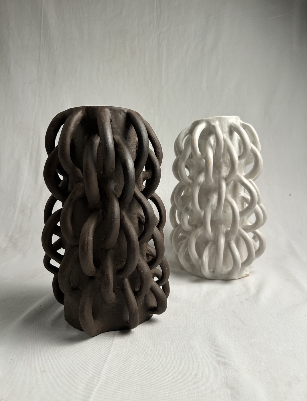 Two hand-built ceramic vessels formed from interlocking clay loops, one dark stoneware and one white, by Consuelo Becerra