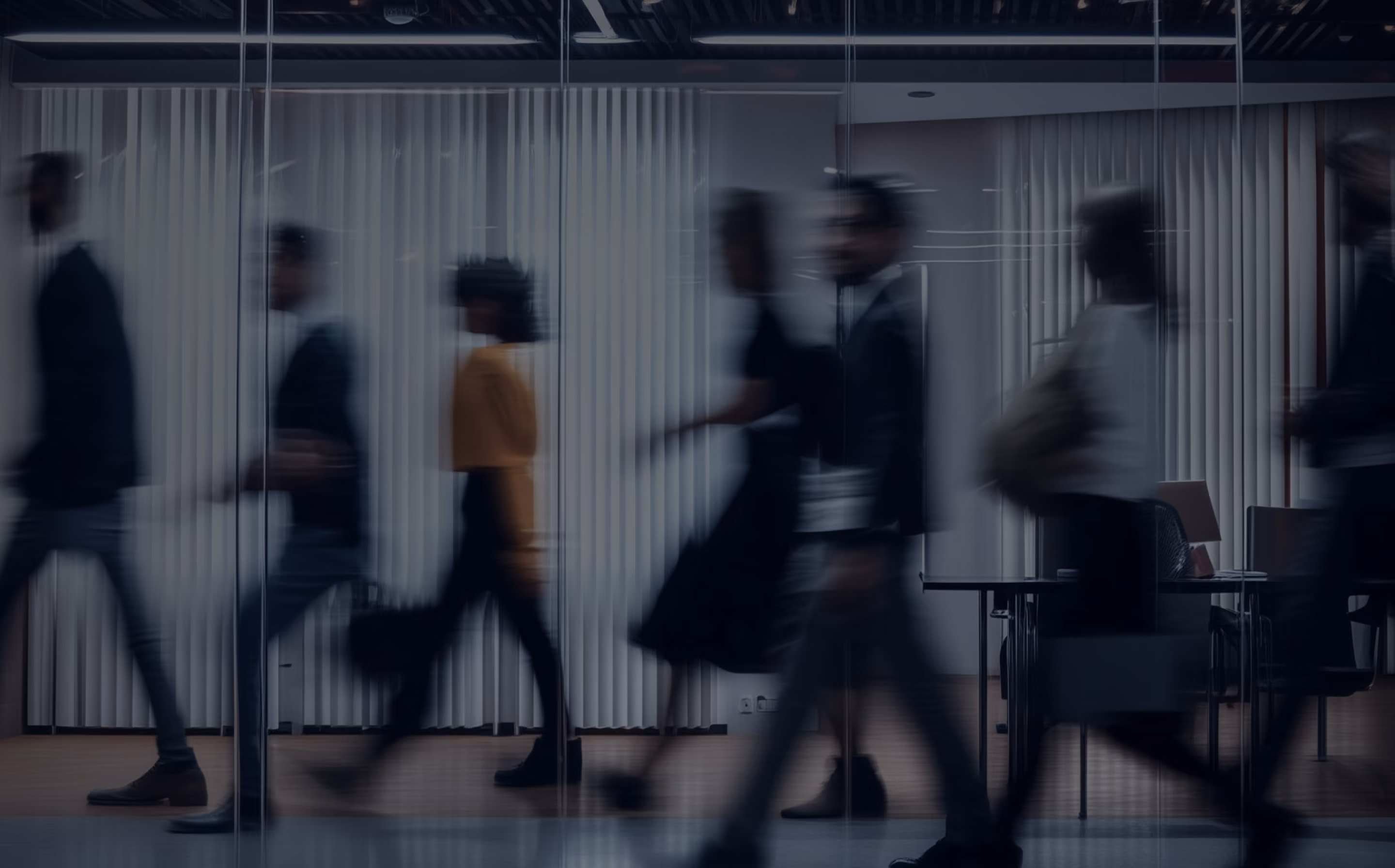 Blurred silhouettes of people walking briskly inside a modern office with glass walls and white vertical blinds.