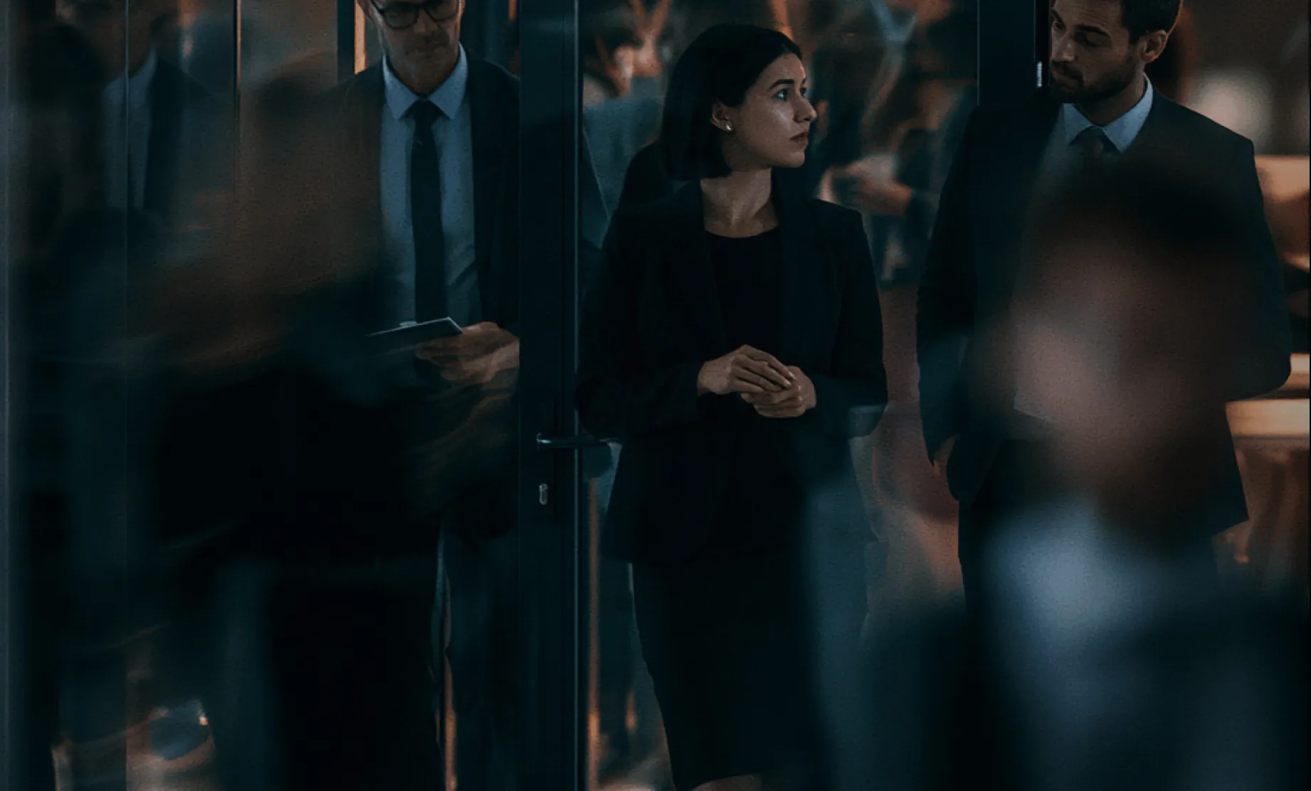 A businesswoman in a dark suit standing between two businessmen in suits in an office setting with blurred figures in the foreground.