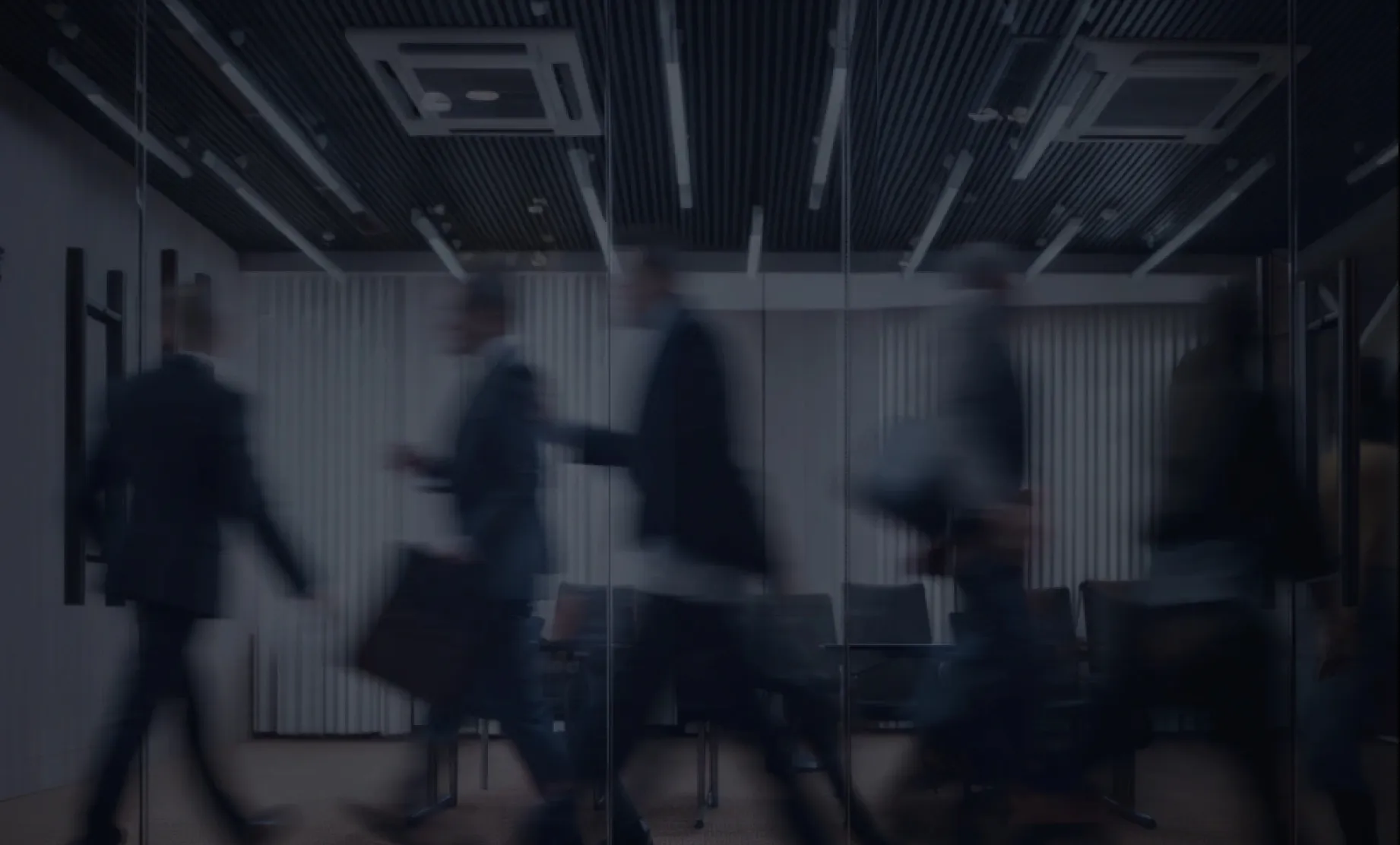 Blurred silhouettes of businesspeople walking past glass walls in a modern office space with chairs and ceiling lights.