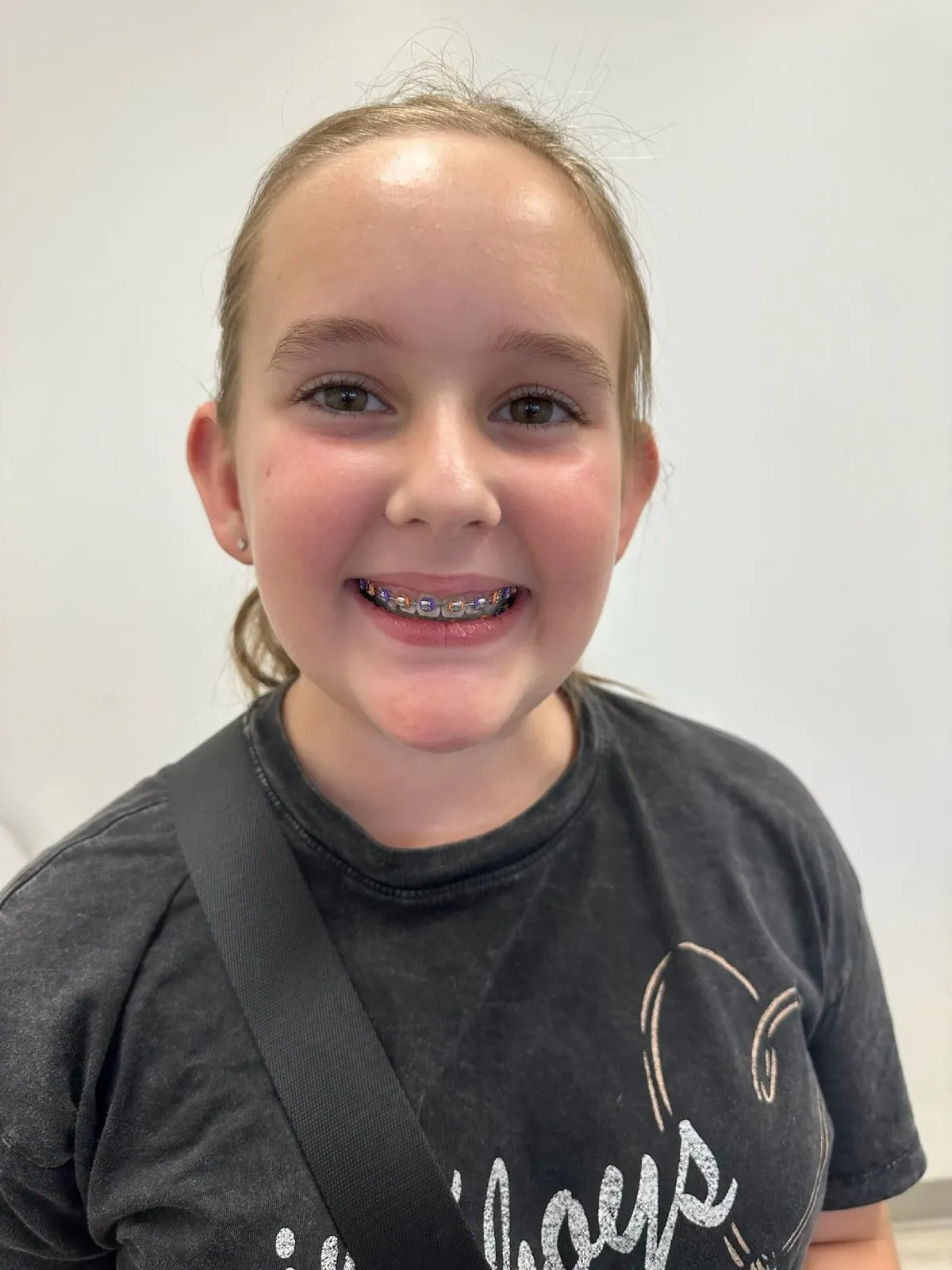 Smiling young girl with braces, flushed cheeks, and hair pulled back, wearing a black T-shirt and shoulder strap against a plain wall.