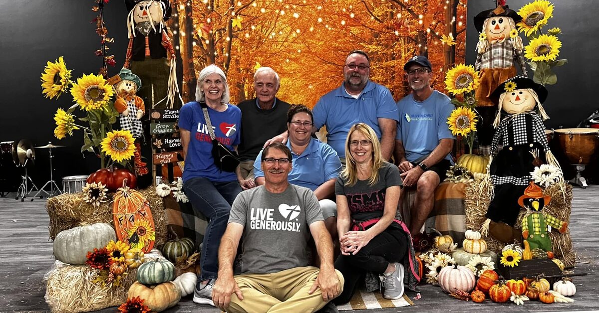 Members at Alive in Christ Lutheran Church in Marana, Arizona pose together at a fall festival. They are surrounded by hay bales, pumpkins, and scarecrows all sitting in front of an orange background.  