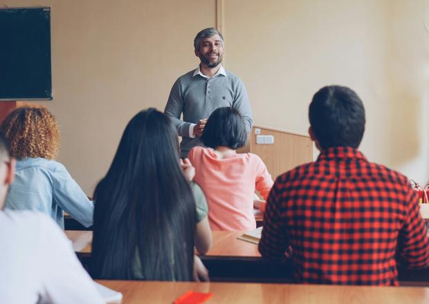 A stock photo of a teacher lecturing a classroom. The teacher is facing the camera, which is behind the students