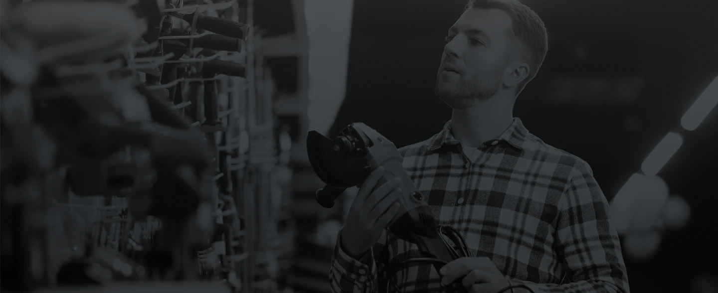 Man in plaid shirt examining an electric angle grinder in a tool store aisle.