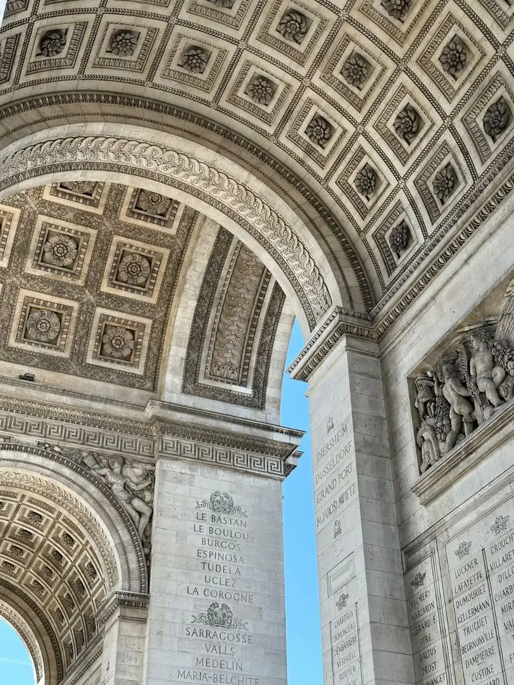 Détail du plafond sculpté de l'Arc de Triomphe à Paris montrant des motifs floraux en relief dans des caissons carrés et des inscriptions de noms de villes gravées sur les murs en pierre claire.