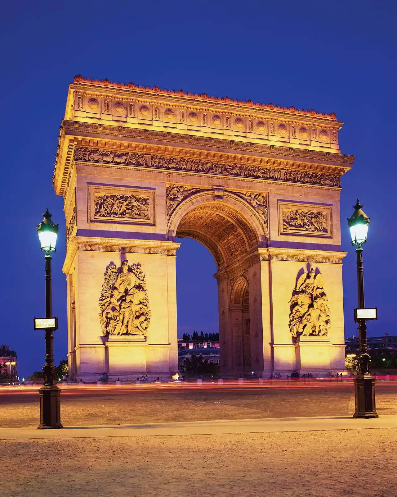 Vue de nuit de l'Arc de Triomphe à Paris, éclairé en jaune, avec deux lampadaires allumés de chaque côté et un ciel bleu foncé en arrière-plan.