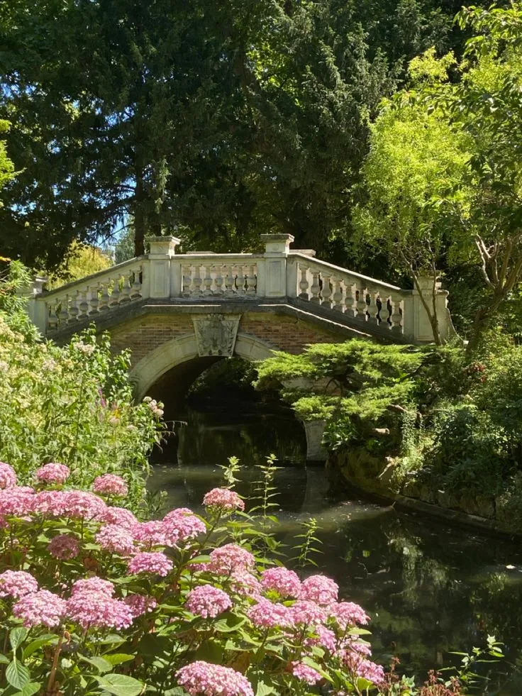 Un pont en pierre orné avec des balustrades, traversant un cours d'eau bordé de fleurs roses et de végétation luxuriante sous un ciel bleu visible à travers les arbres.