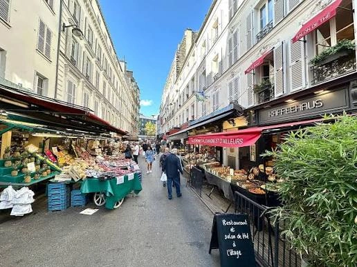 Rue piétonne bordée de bâtiments traditionnels avec des stands de fruits, légumes et pâtisseries à gauche et un café-restaurant à droite sous un ciel bleu clair.