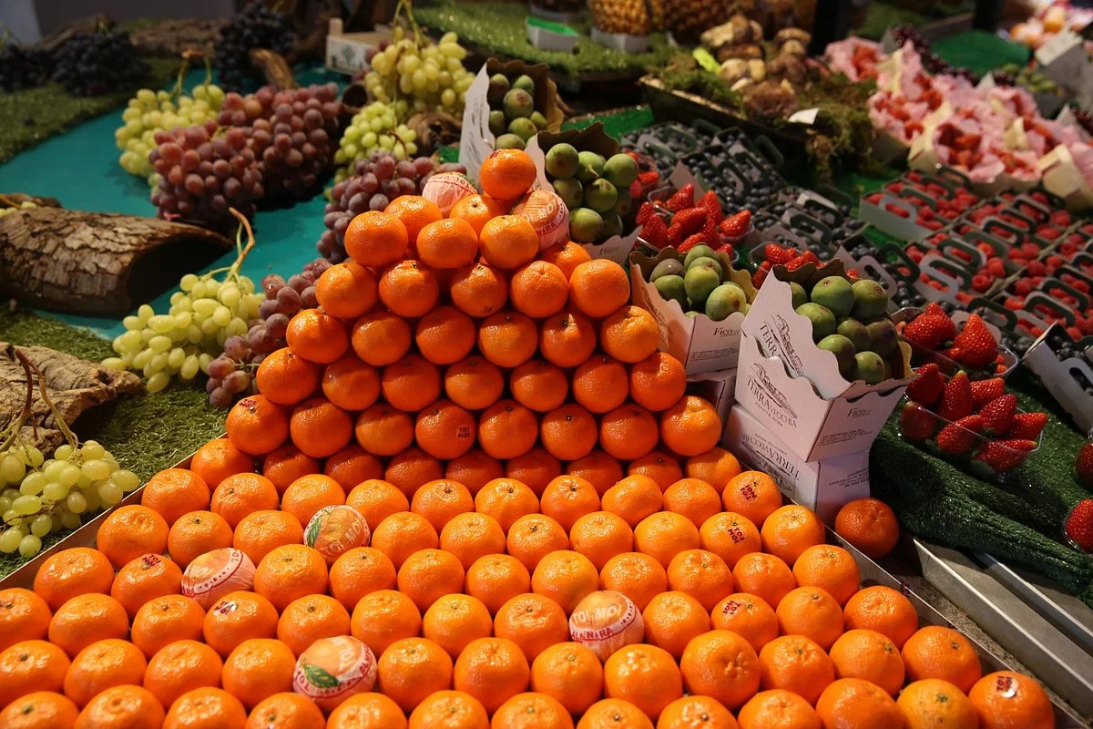 Un étalage de marché avec une pyramide de mandarines orange vif au centre, entourée de grappes de raisins verts et rouges, de fraises et de caisses de figues vertes, sur une table recouverte de gazon synthétique.