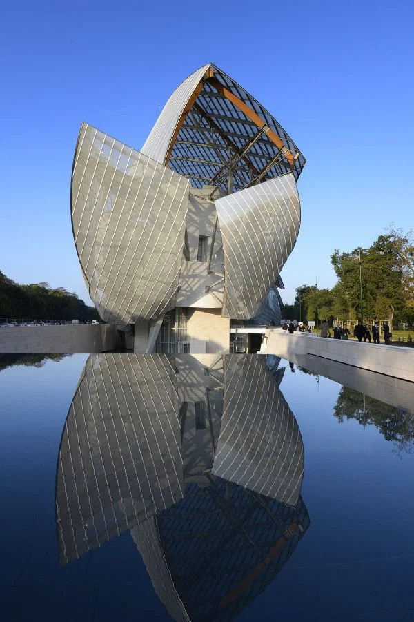 Le bâtiment de la Fondation Louis Vuitton avec sa structure métallique et vitrée en forme de voiles, reflété dans une grande étendue d'eau calme sous un ciel bleu clair.
