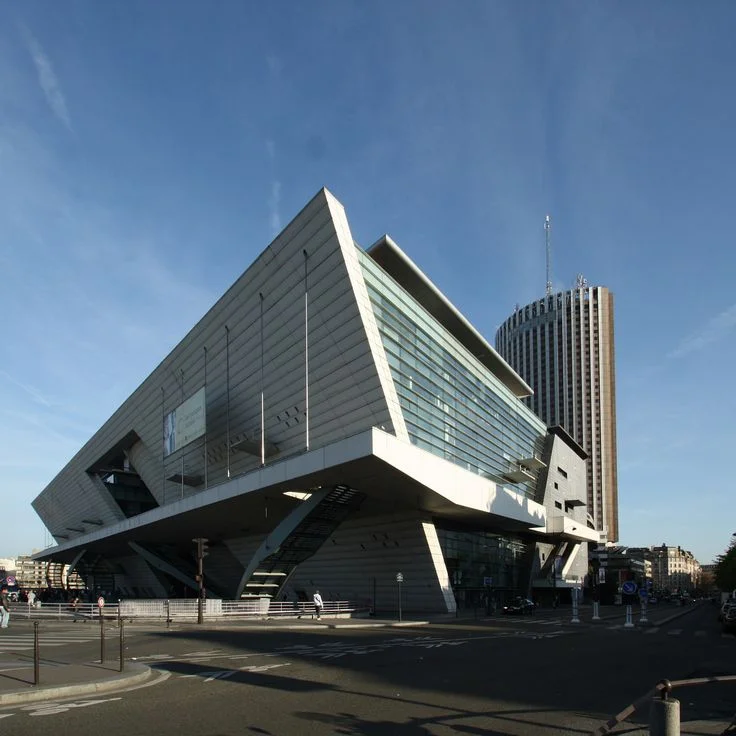 Bâtiment moderne en forme triangulaire avec des murs en verre et métal sous un ciel bleu, avec une tour cylindrique en arrière-plan.