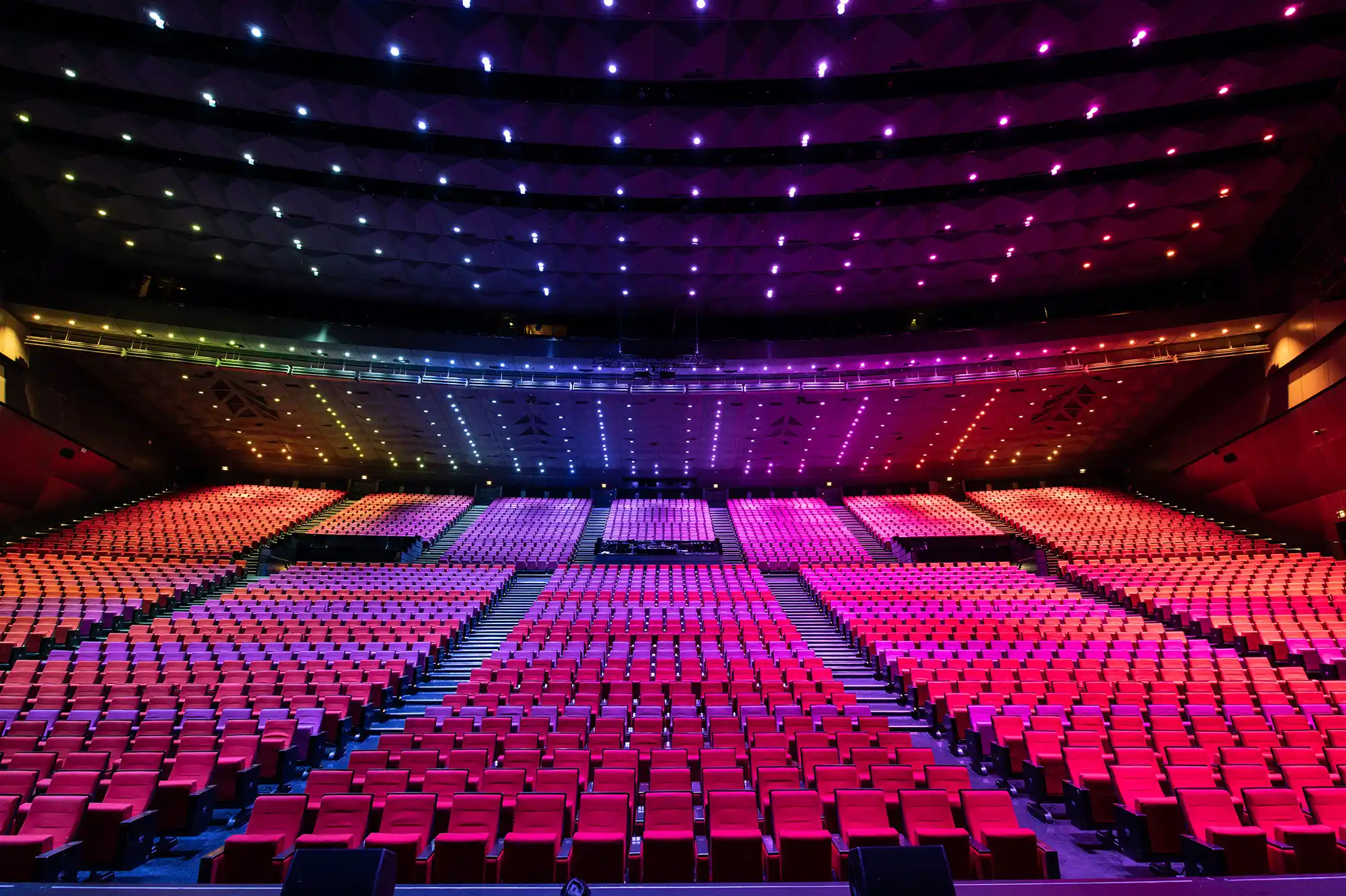 Intérieur d'un grand auditorium avec des rangées de sièges rouges organisées en sections divisées par des allées, éclairé par des lumières colorées au plafond variant du violet au rouge.