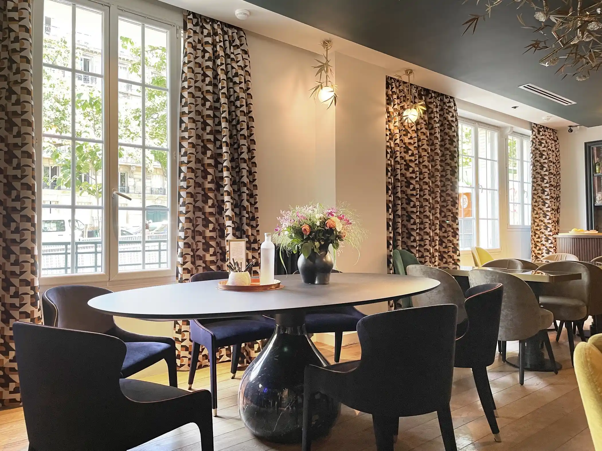 Sala de jantar iluminada com grandes janelas emolduradas por cortinas com padrão geométrico em tons de castanho e bege, mesa oval preta com base em forma de vaso rodeada por cadeiras modernas pretas e azuis, decorada com um ramo de flores colorido e um jarro branco.