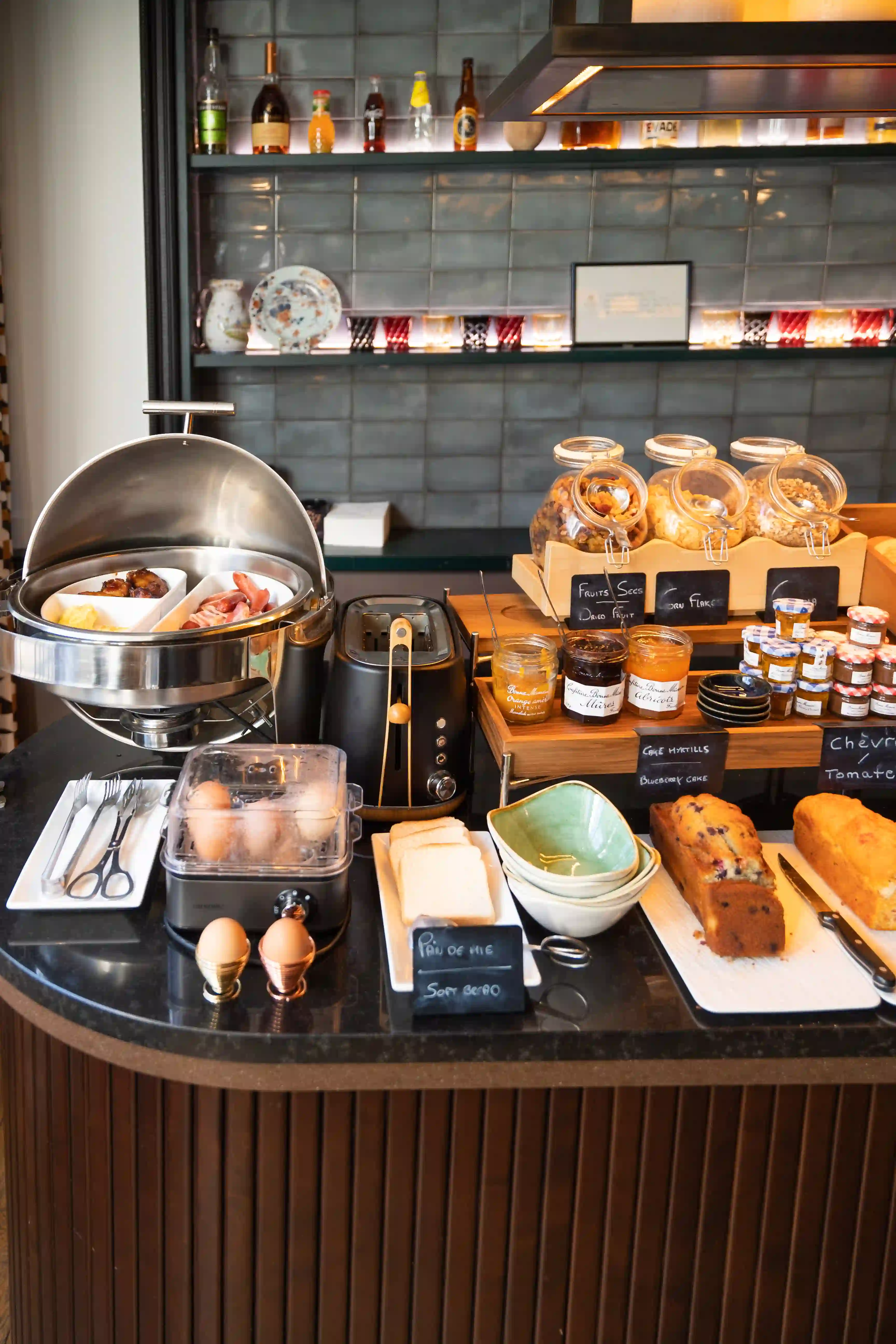 Buffet de petit-déjeuner avec cuiseur à œufs contenant des œufs, grille-pain noir, plateau avec beignets, bacon, et œufs brouillés, bocaux de fruits secs, flocons d'avoine, pots de confiture variée, tranches de pain, bols empilés, et deux cakes, le tout sur un comptoir noir devant un mur carrelé gris avec étagères décorées.