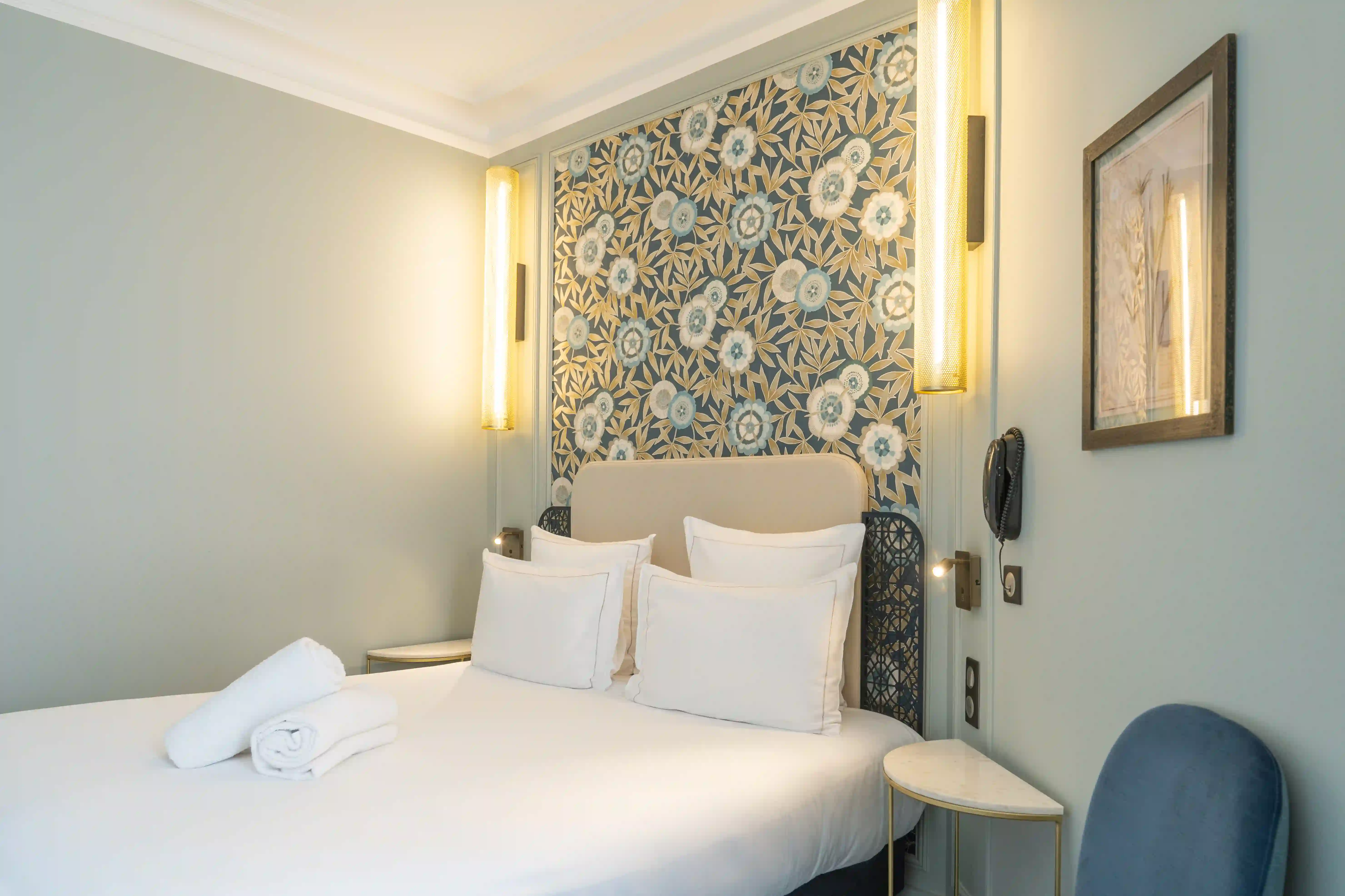 Chambre avec lit double blanc, quatre oreillers alignés, et deux serviettes blanches pliées dessus. Tête de lit beige avec panneau floral bleu, beige et blanc derrière. Deux lampes murales jaunes allumées de chaque côté, deux tables de chevet blanches avec pieds dorés, chaise bleue et cadre avec dessin de plantes sur mur bleu clair.