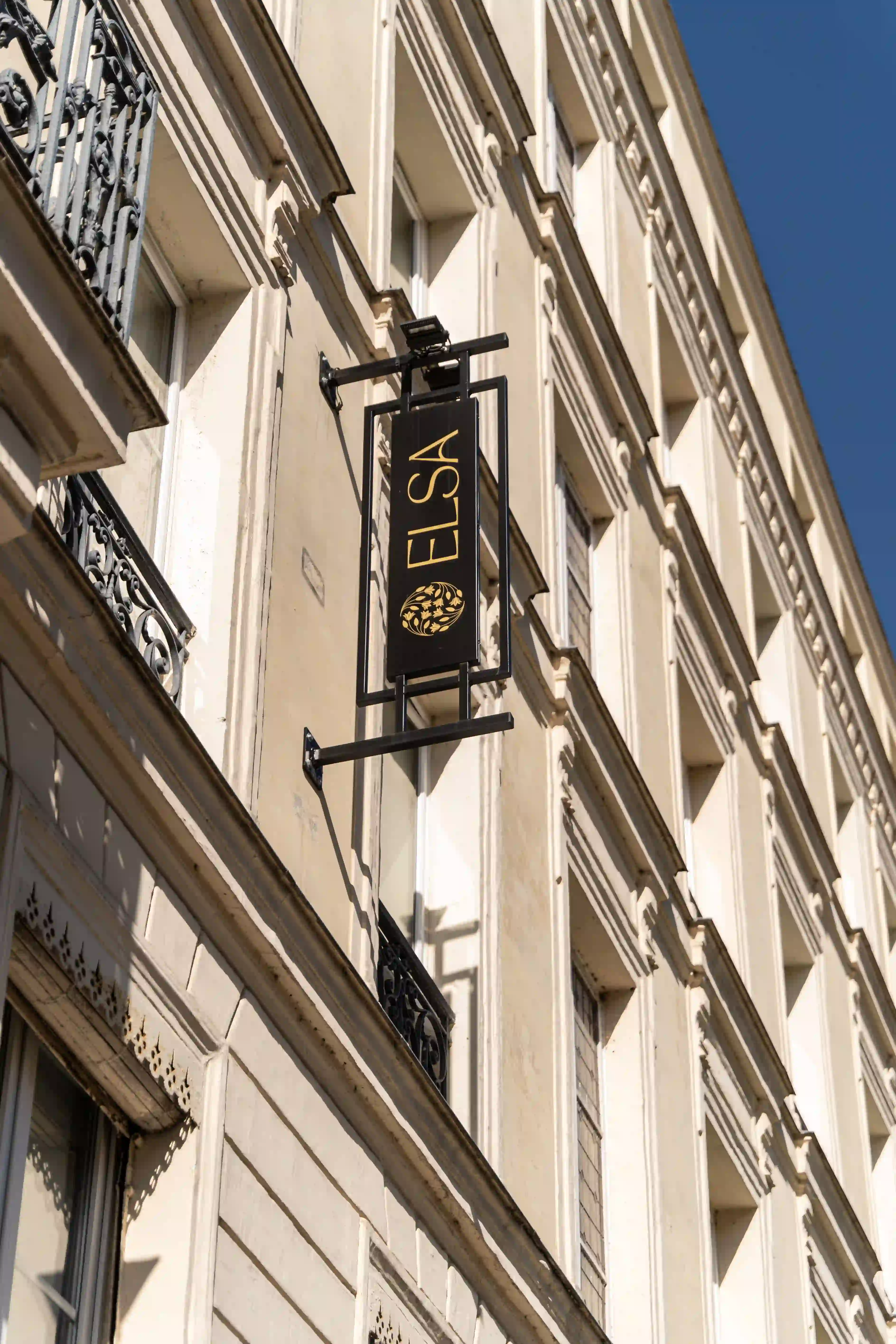 Façade d'un bâtiment beige avec balcons en fer forgé et un panneau suspendu noir avec l'inscription dorée « ELSA » et un logo circulaire décoratif, sous un ciel bleu clair.
