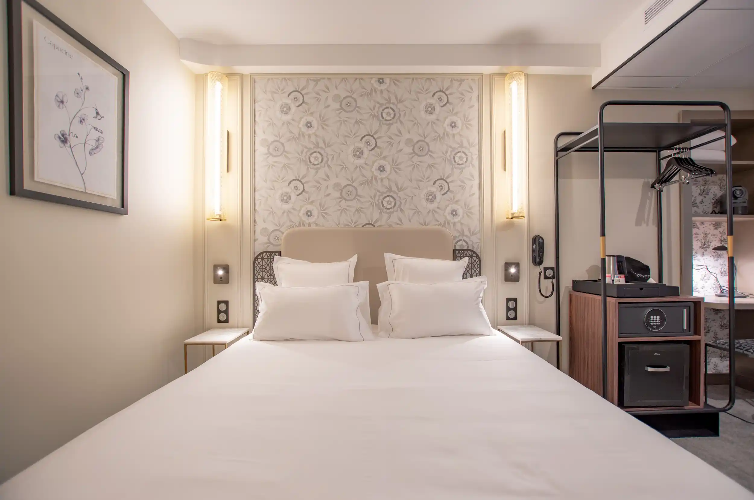 Modernes Hotelzimmer mit einem großen Bett mit beigem Kopfteil, vier weißen Kissen, Wänden mit vertikaler Beleuchtung und floraler Dekoration im Hintergrund, einem kleinen Nachttisch auf jeder Seite sowie einem Möbelstück mit Safe und Minikühlschrank auf der rechten Seite.