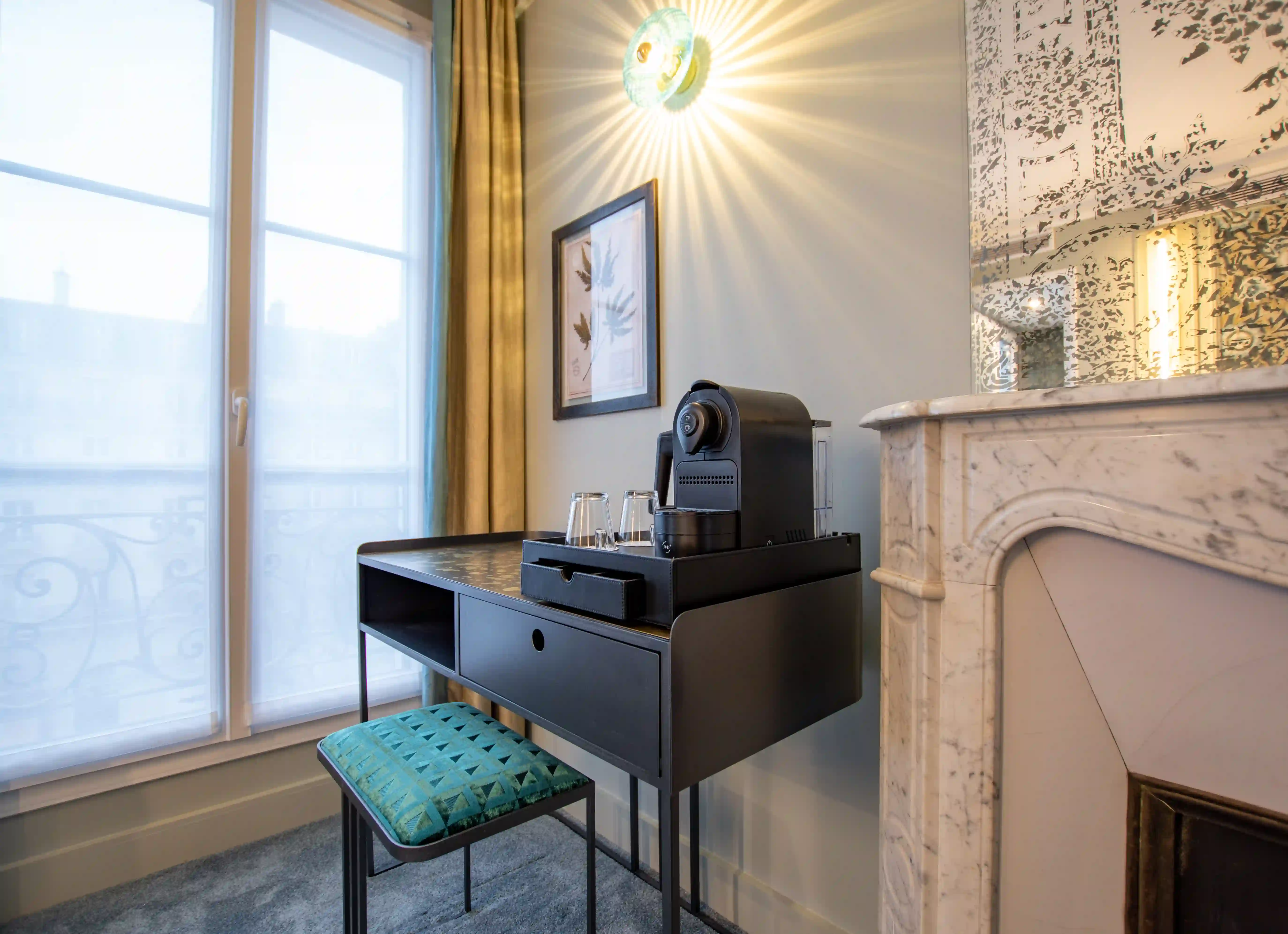 Coin bureau moderne noir avec plateau contenant une machine à café et deux verres, tabouret tapissé bleu canard, grande fenêtre avec voilages, tableau mural, applique lumineuse rayonnante, et cheminée en marbre blanc avec miroir ancien.