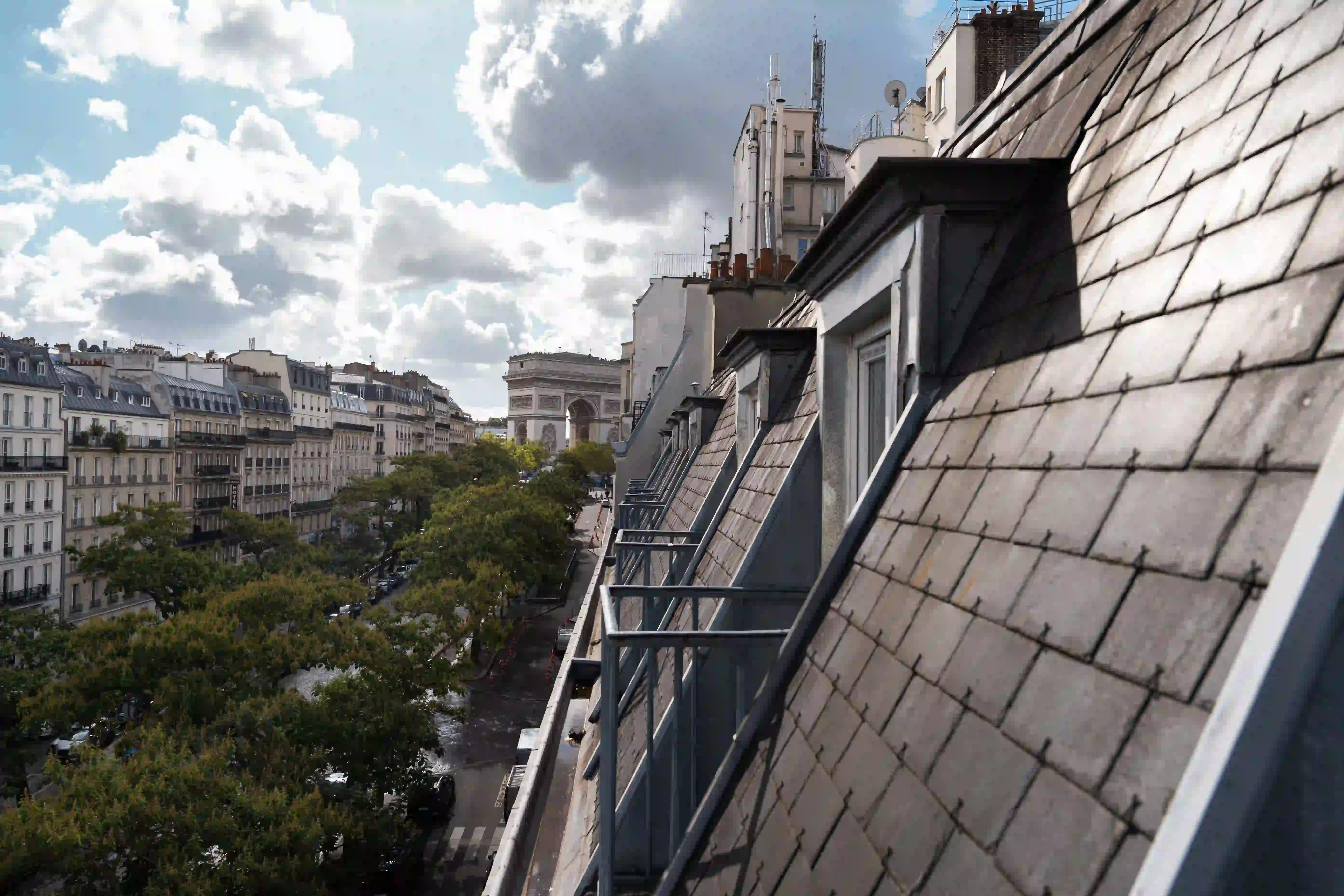 Vue inclinée sur des toits d'immeubles parisiens avec des balcons en fer, une avenue bordée d'arbres et l'Arc de Triomphe au loin sous un ciel partiellement nuageux.