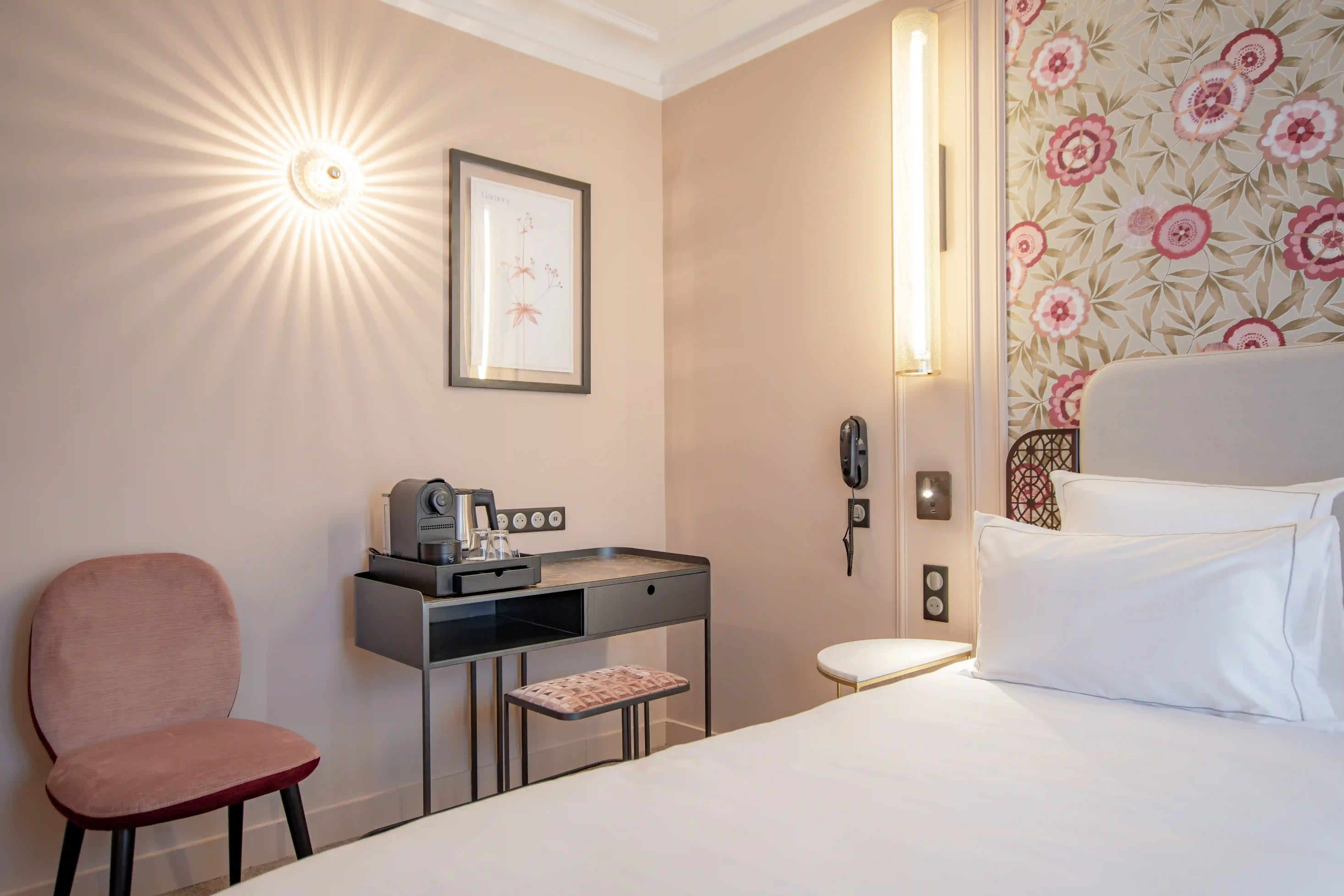 Chambre d'hôtel élégante avec un lit à tête de lit blanche, une lampe murale allumée, un bureau noir avec une machine à café, deux verres et un tabouret en tissu rosé, une chaise rose et un mur à motif floral rose et beige.