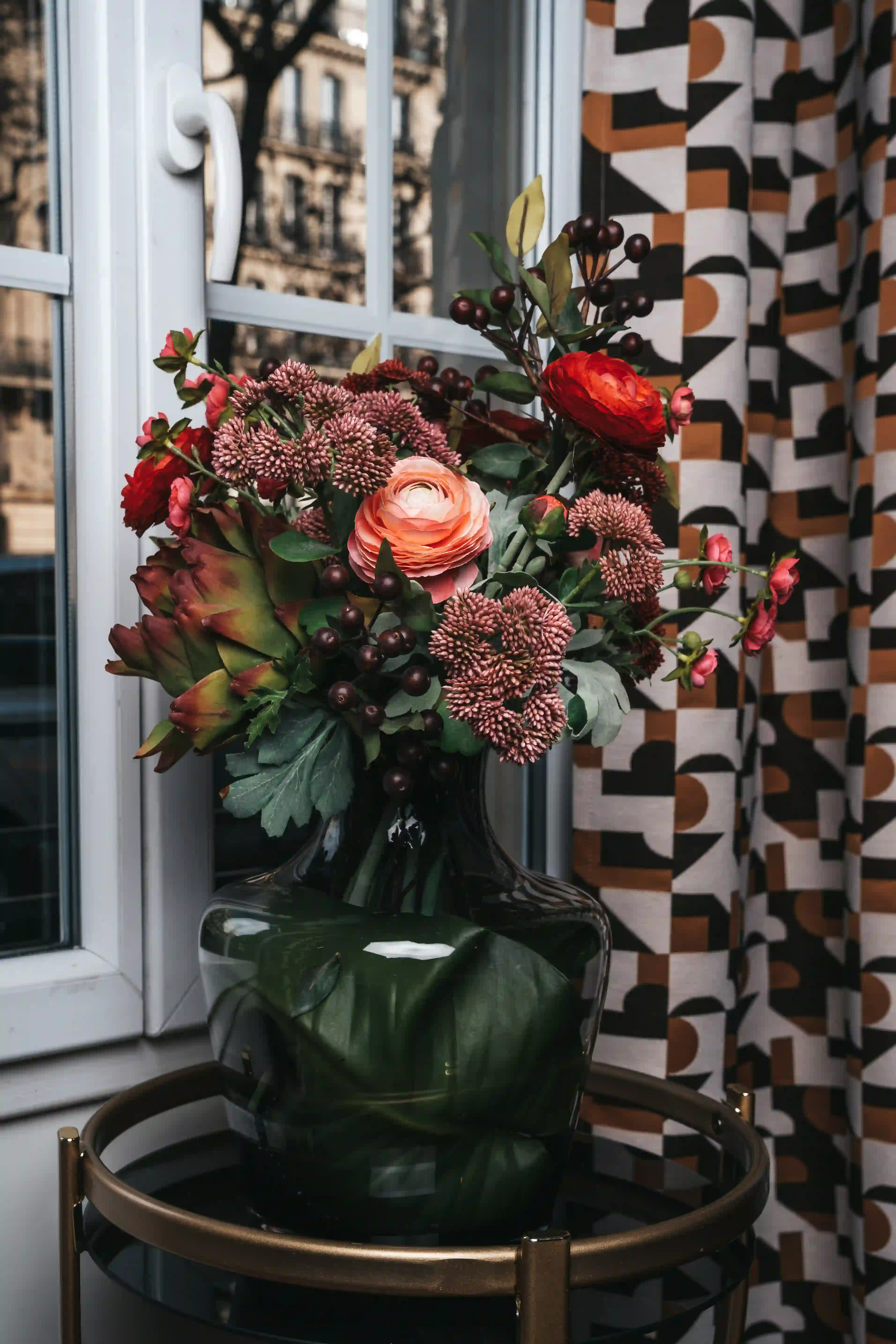 Bouquet of colorful flowers including coral roses, red flowers, dark berries, and green foliage, in a dark glass vase placed on a round table in front of a window with an orange, black, and white geometric curtain.