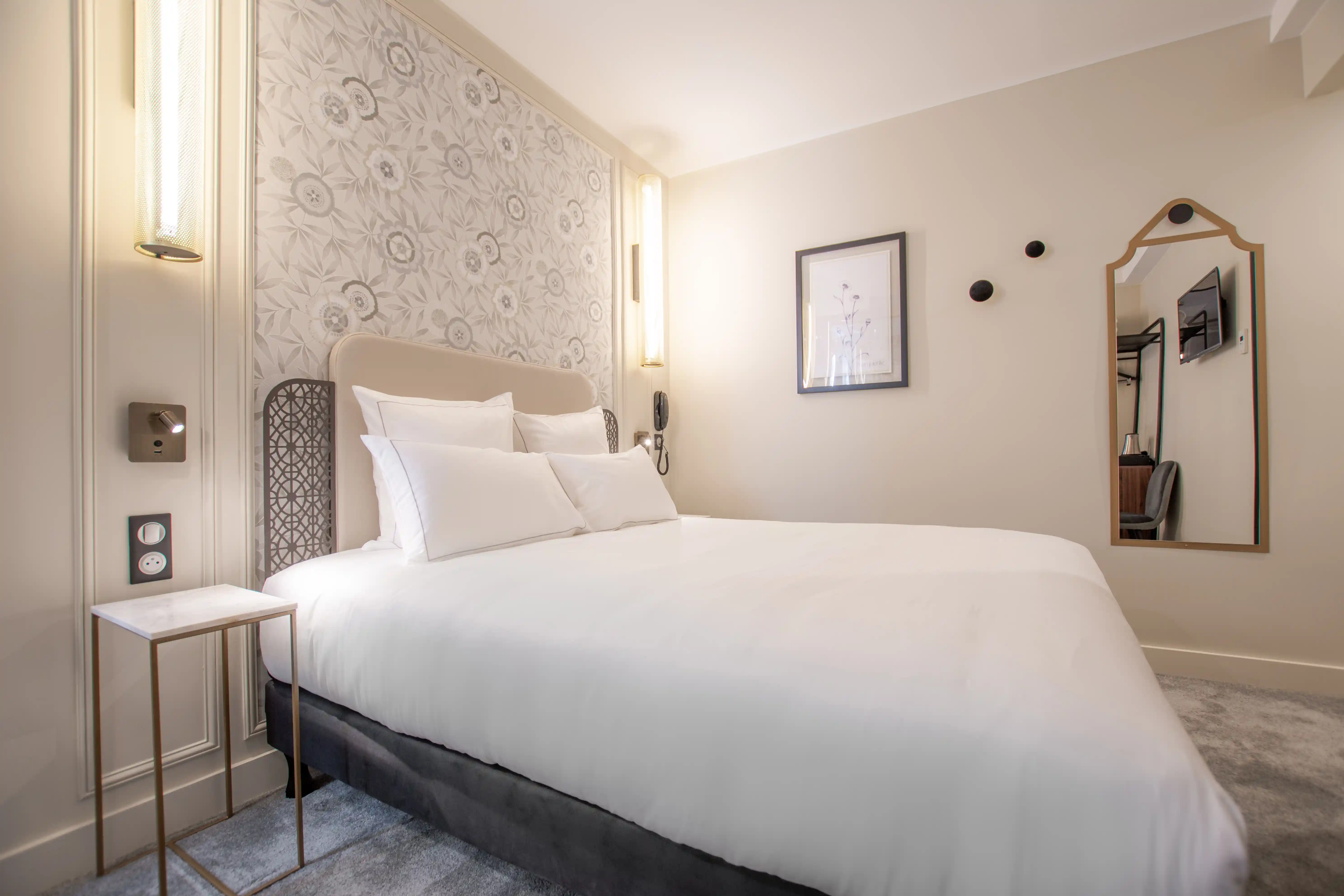 Chambre d'hôtel élégante avec un grand lit blanc, une tête de lit beige et un mur décoré d'un papier peint à motifs floraux gris. À gauche, une petite table d'appoint avec un plateau blanc et une lampe murale allumée. Au mur de droite, un cadre avec une illustration florale, un miroir mural avec cadre en bois clair et des patères noires. Le sol est recouvert d'un tapis gris clair.