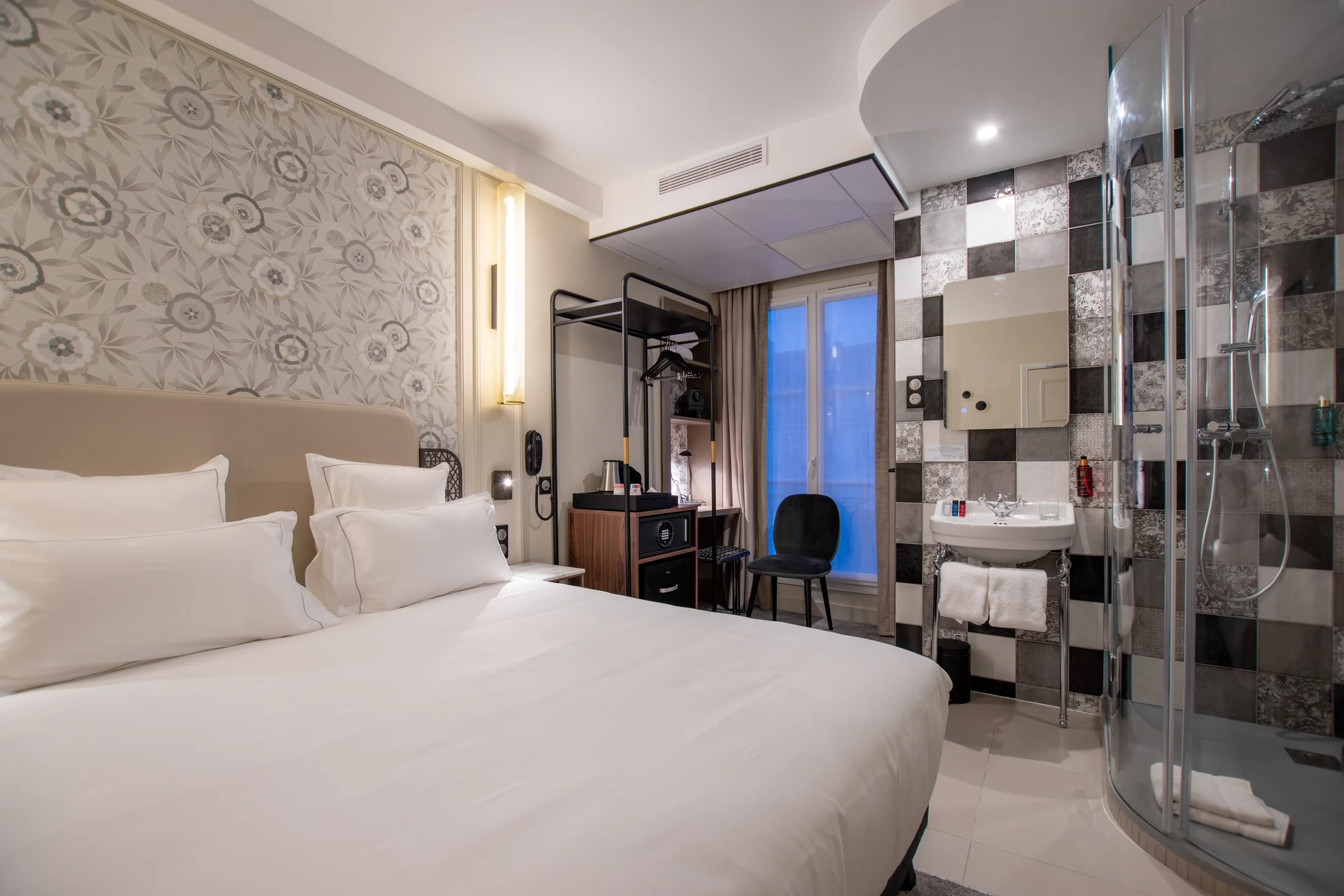 Chambre d'hôtel moderne avec grand lit double blanc, tête de lit beige, papier peint floral gris et blanc, porte-manteaux noir, chaise noire, fenêtre avec rideaux, lavabo avec miroir, douche à l'italienne avec paroi en verre et carrelage noir et gris.