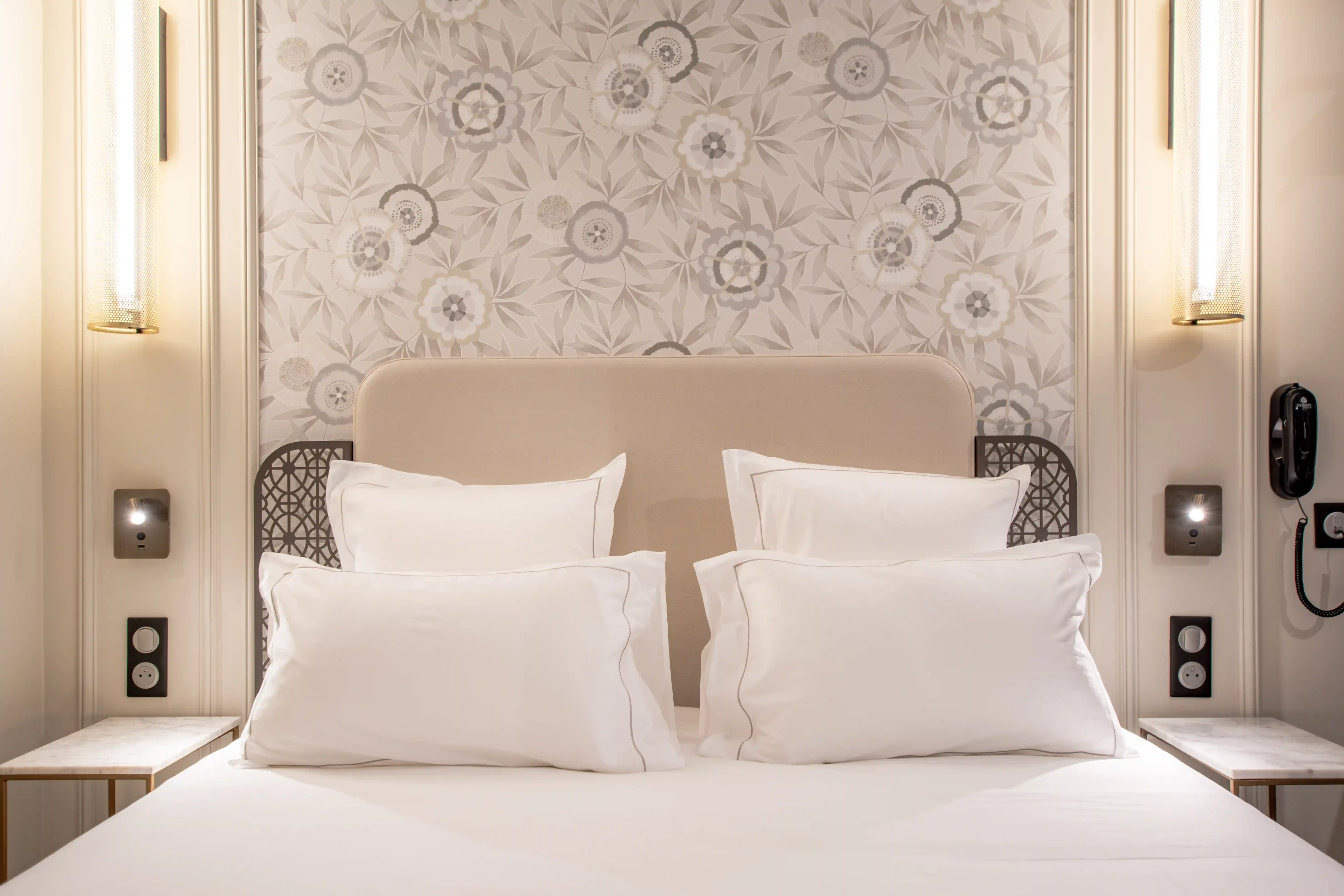 Lit double avec tête de lit beige, quatre oreillers blancs et une tête de lit métallique ajourée, éclairage mural de chaque côté et papier peint floral gris beige en arrière-plan.