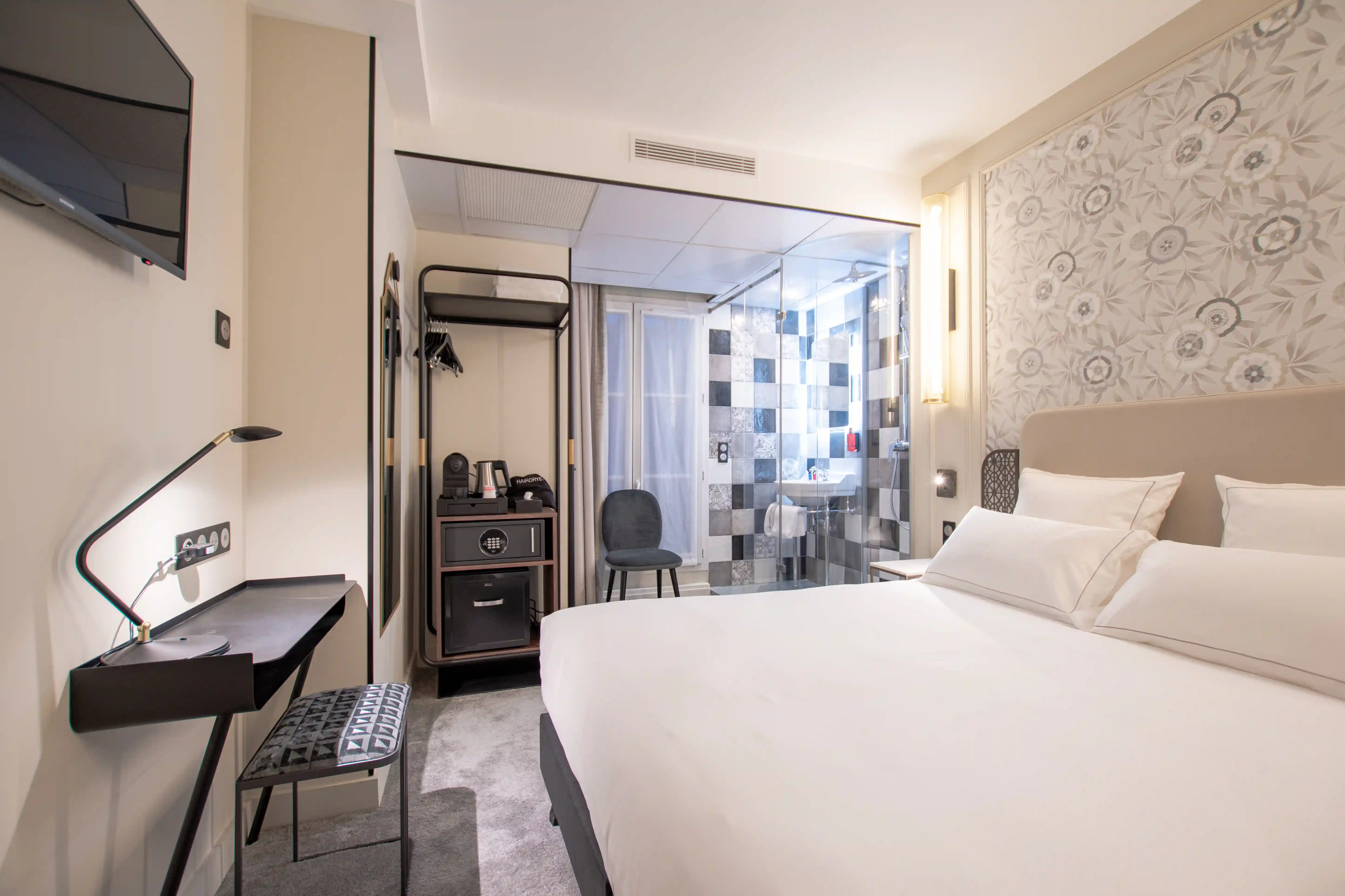 Chambre d'hôtel moderne avec un grand lit double à linge blanc, un mur à motifs floraux, une télévision fixée au mur, un bureau noir avec lampe, un fauteuil, un porte-manteau ouvert et une salle de bain vitrée avec douche à carreaux noirs et gris.