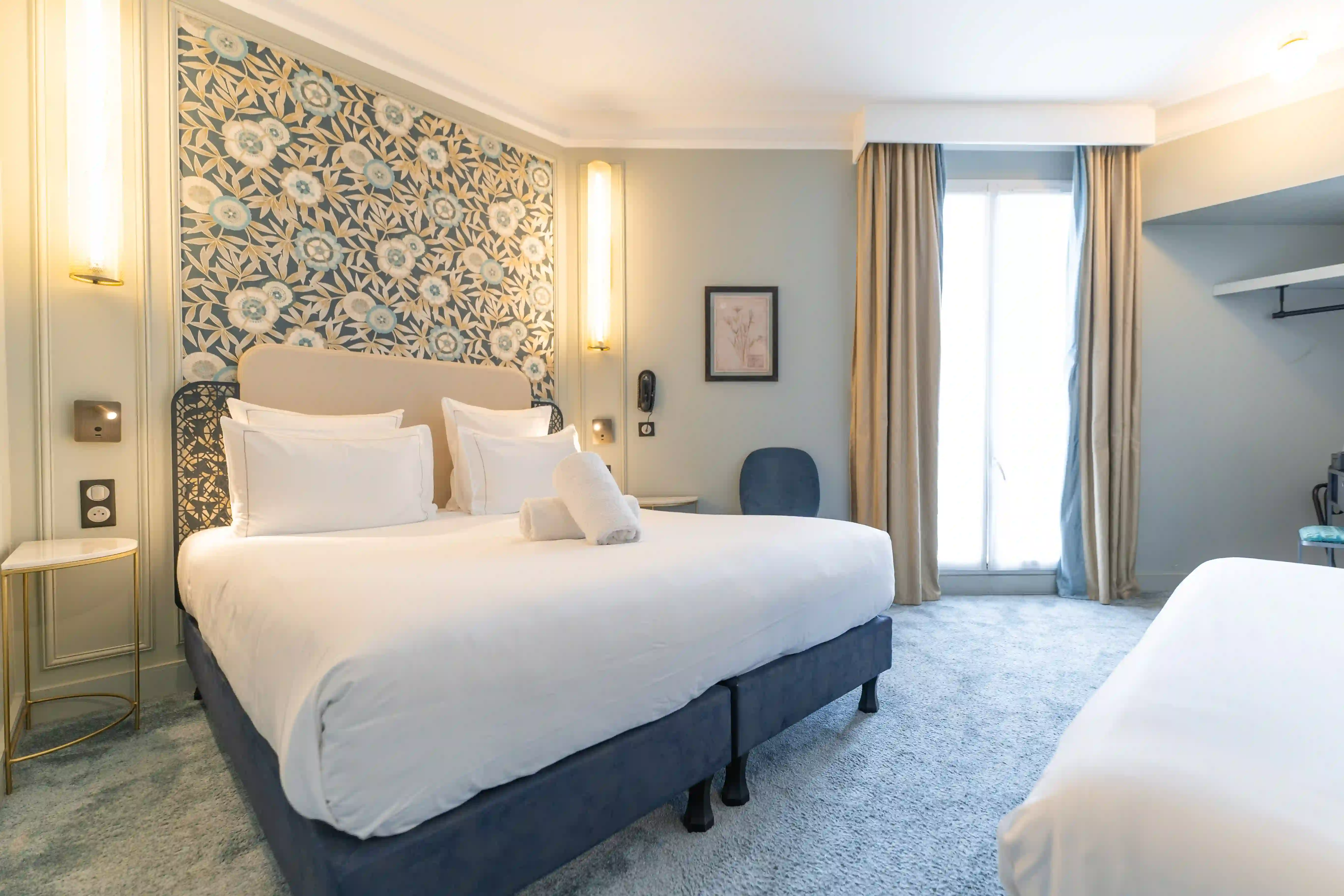 Chambre d'hôtel lumineuse avec un grand lit double à tête de lit beige, papier peint floral bleu et blanc derrière, table de chevet ronde avec lampe murale, chaise bleue, fenêtre avec rideaux beige et moquette grise.