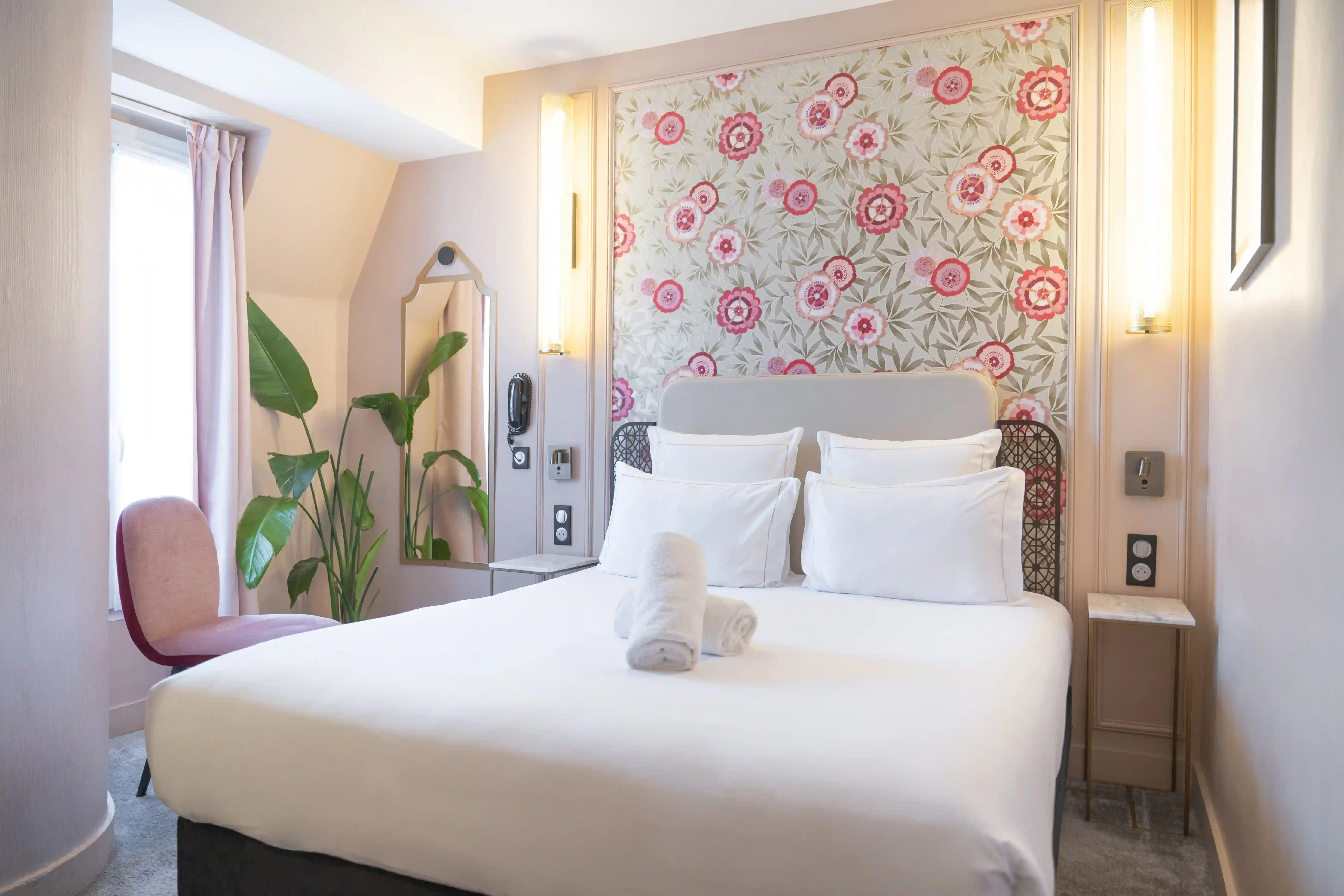 Chambre avec un grand lit double décoré de quatre oreillers blancs et deux serviettes roulées posées dessus, mur avec papier peint floral rose, une plante verte à côté d'une chaise rose près d'une fenêtre, deux appliques murales allumées de chaque côté du lit.