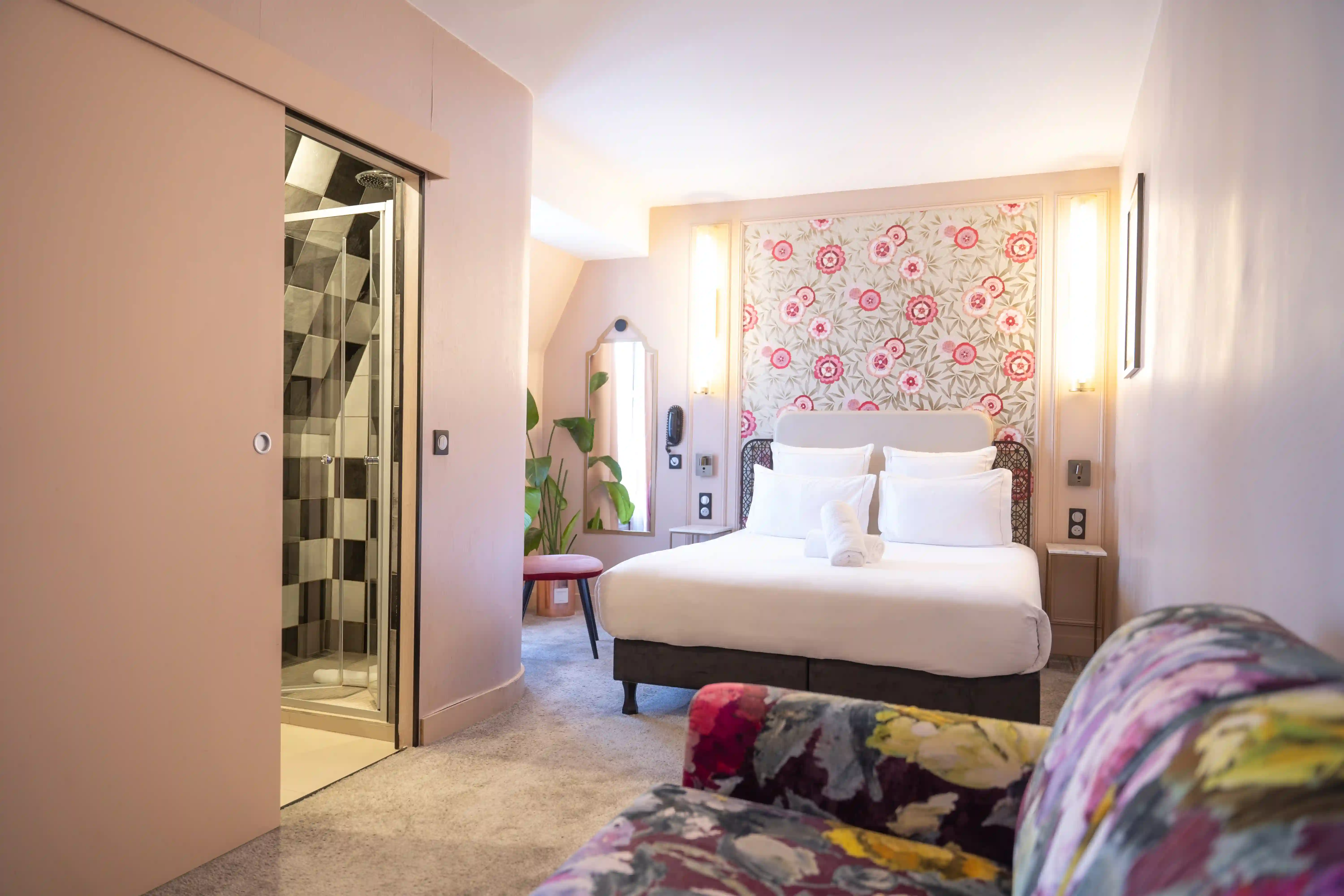 Chambre de suite junior lumineuse avec un grand lit double blanc, un mur décoré de fleurs roses, une douche vitrée à motifs géométriques, un miroir sur pied, une chaise rouge et un canapé floral multicolore en avant-plan.