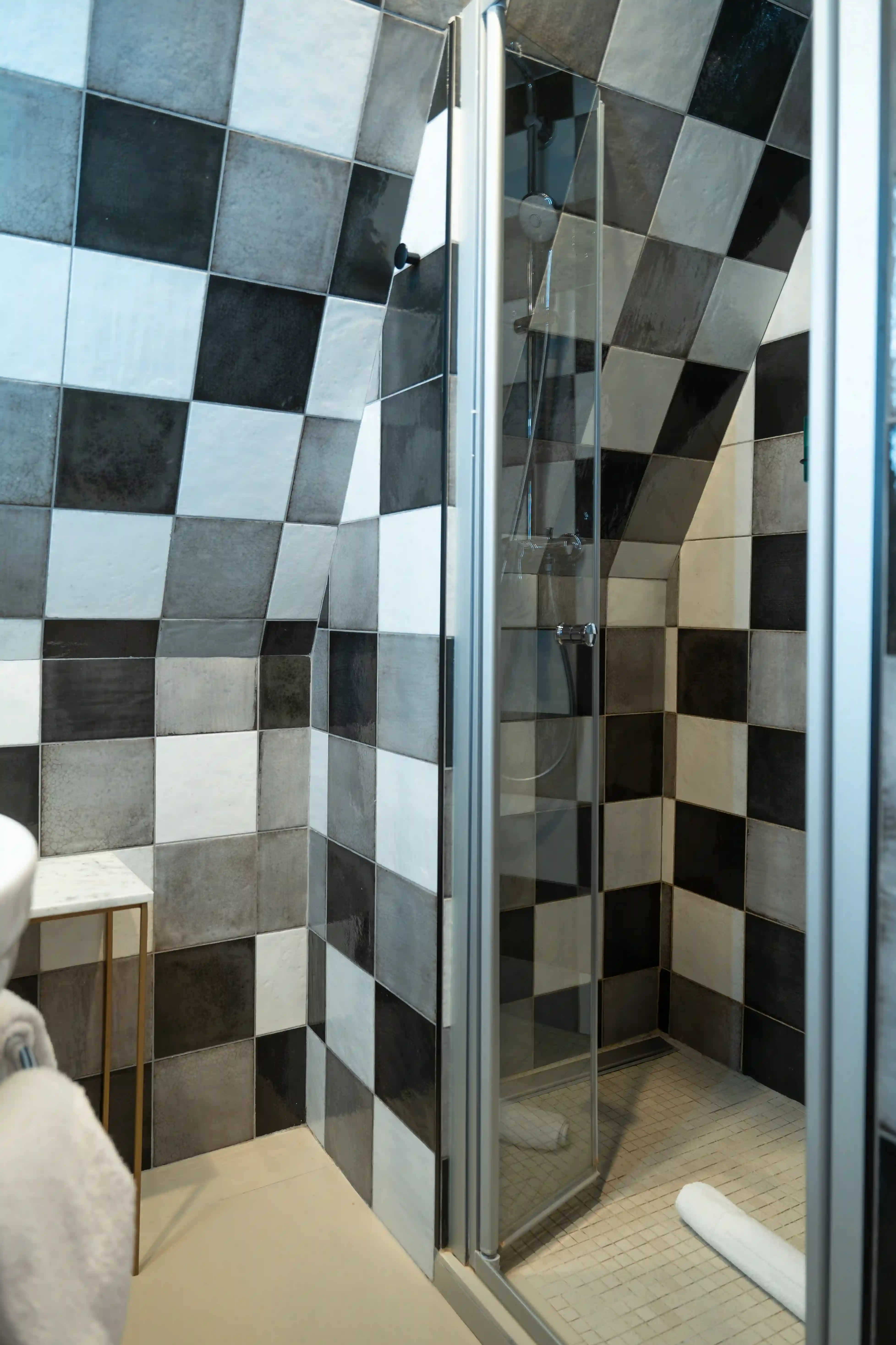 Douche avec parois vitrées et carrelage noir, blanc et gris en damier couvrant les murs et le plafond incliné, avec un sol beige clair et une serviette blanche roulée posée à l'intérieur.