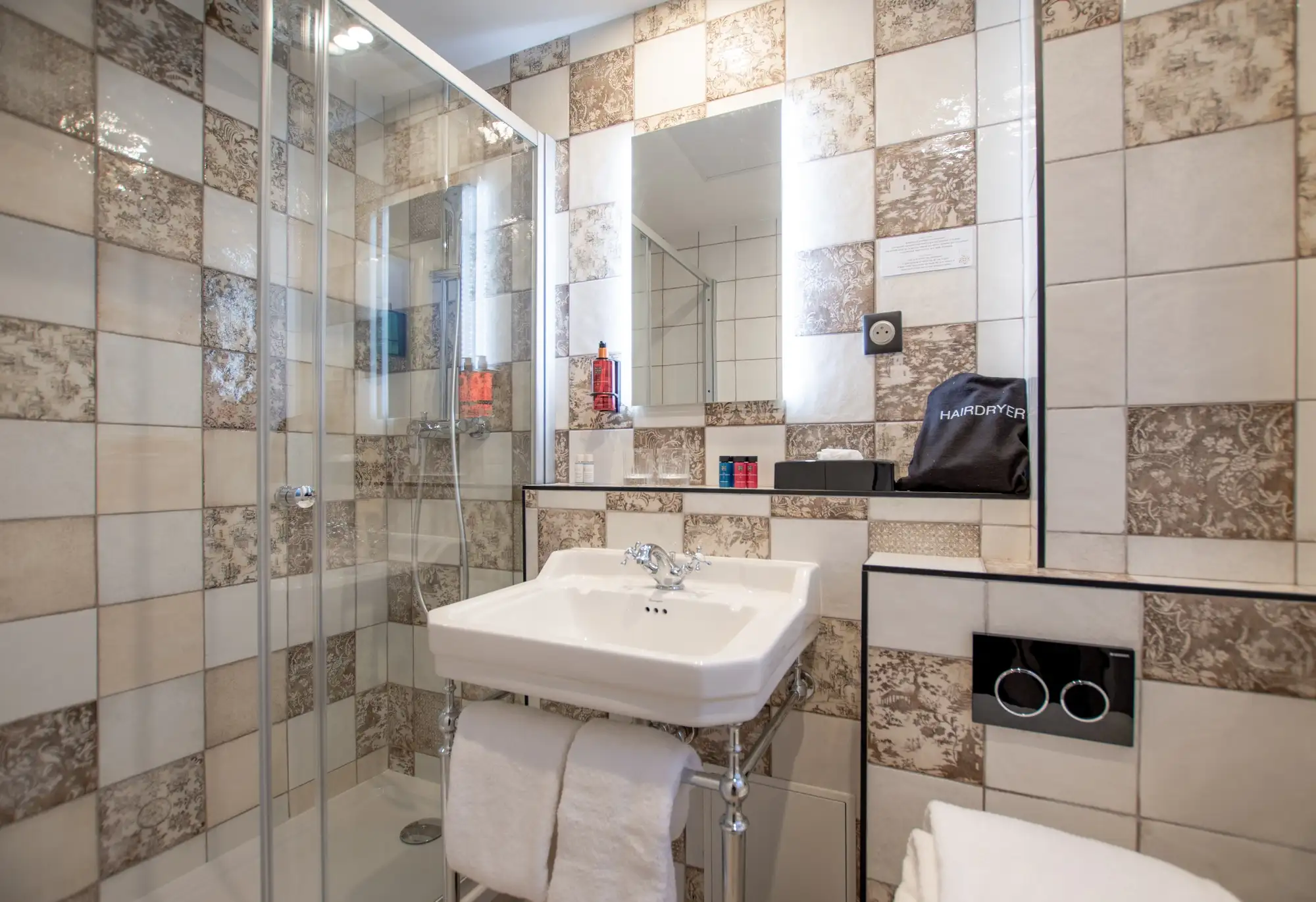 Salle de bain avec douche en verre, lavabo blanc avec serviettes, carreaux beige et marron à motifs floraux, miroir rétroéclairé, sac noir marqué "Hairdryer" et divers articles de toilette.