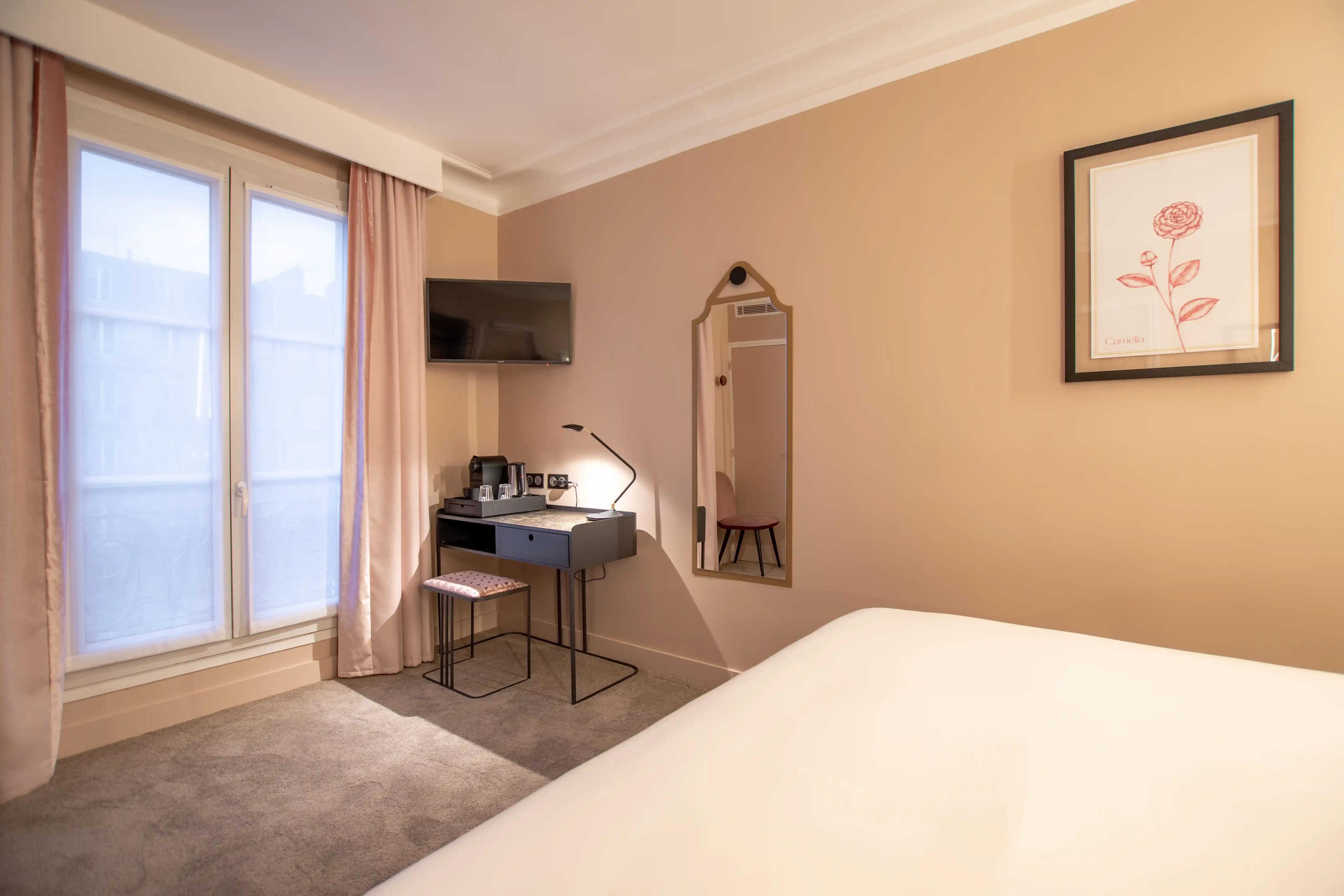 Chambre d'hôtel avec mur beige, fenêtre avec rideaux rose pâle, bureau noir avec lampe, machine à café et verre, chaise rose, miroir mural au cadre décoratif et téléviseur fixé au mur.
