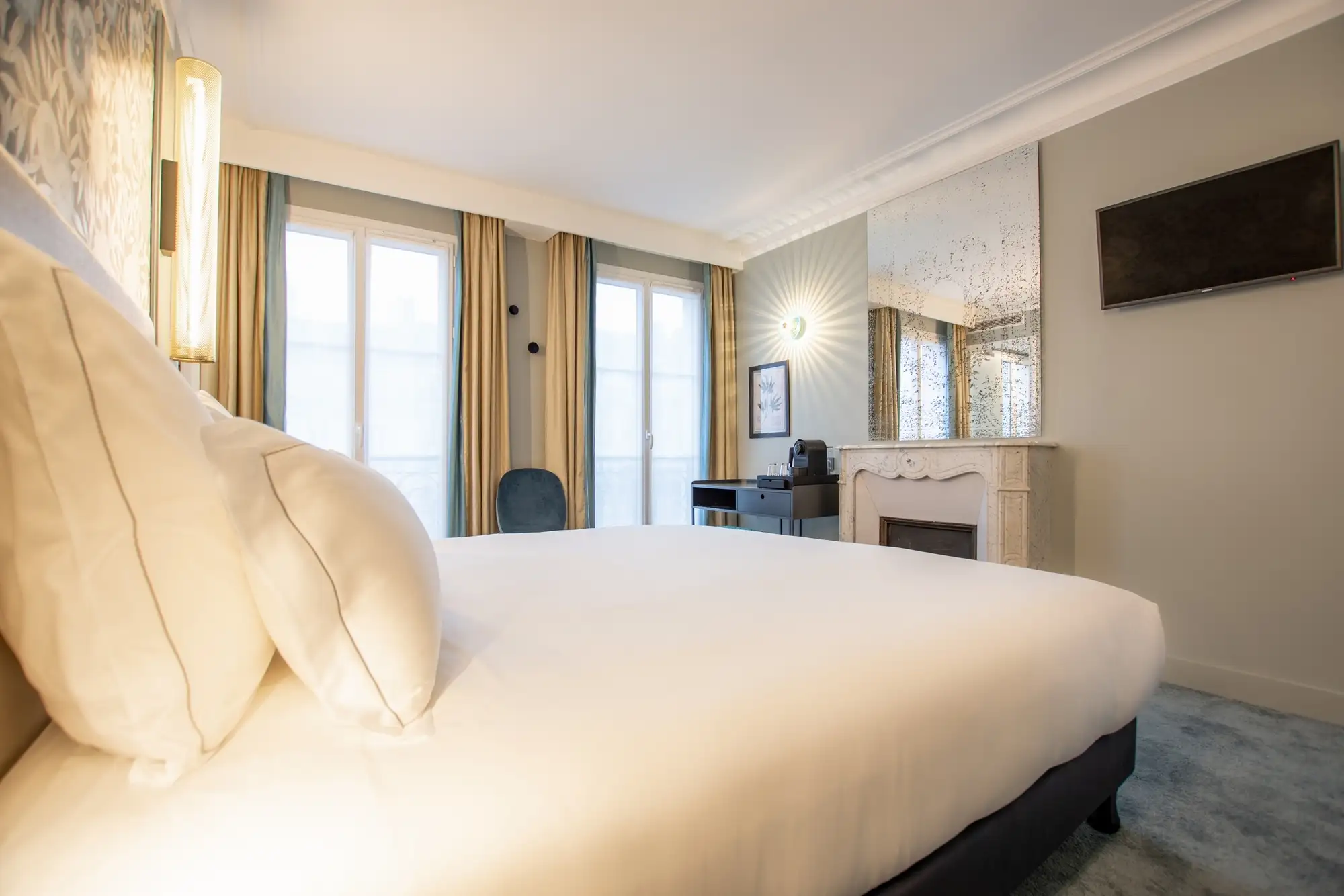 Chambre d'hôtel élégante avec grand lit blanc, cheminée décorative avec grand miroir ancien, télévision murale, bureau noir avec machine à café, et fenêtres avec rideaux beiges et bleus.
