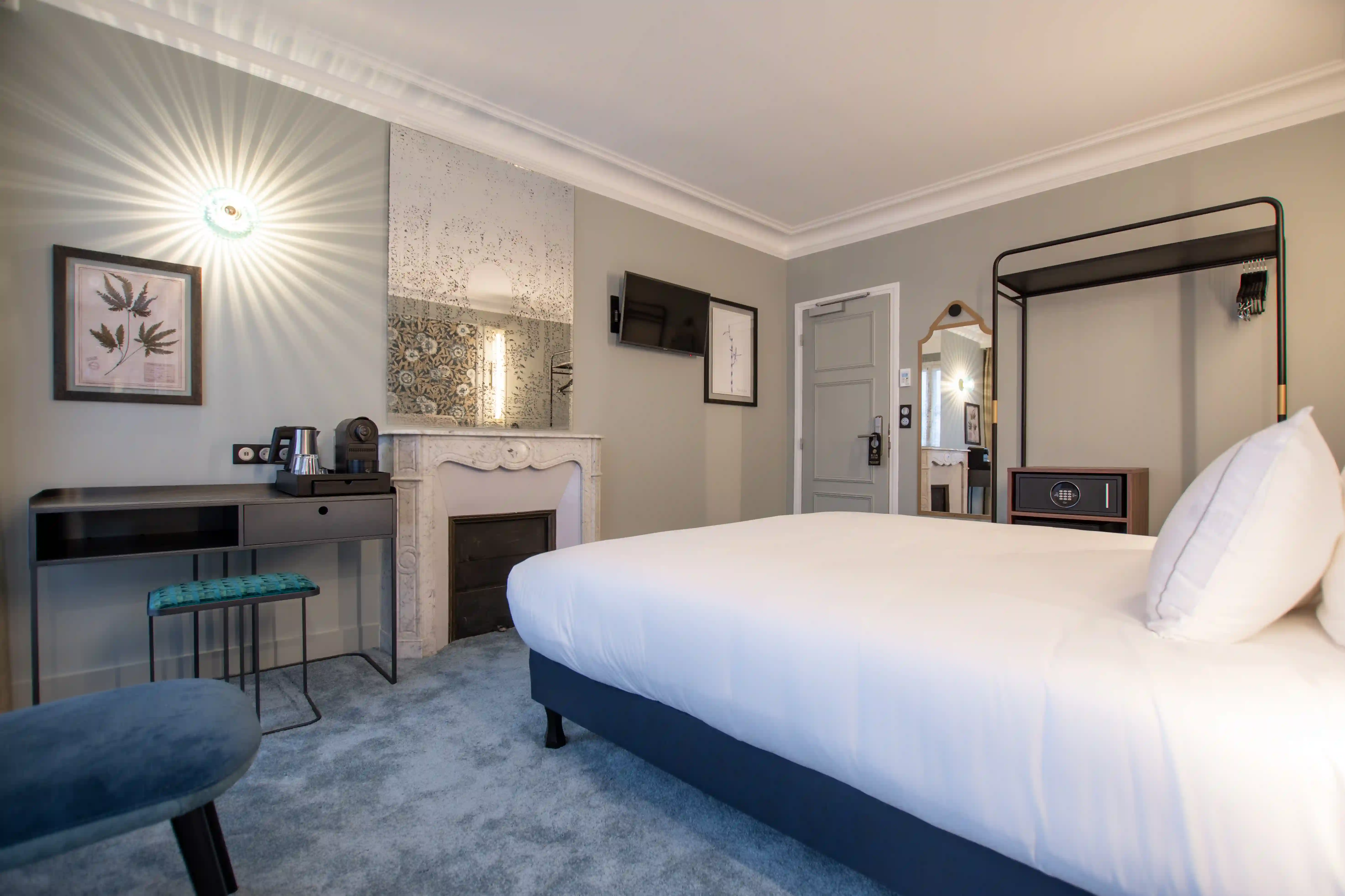 Chambre d'hôtel moderne avec un grand lit blanc, cheminée décorative en marbre, télévision murale, bureau avec machine à café, chaise bleue, miroir et porte grise.