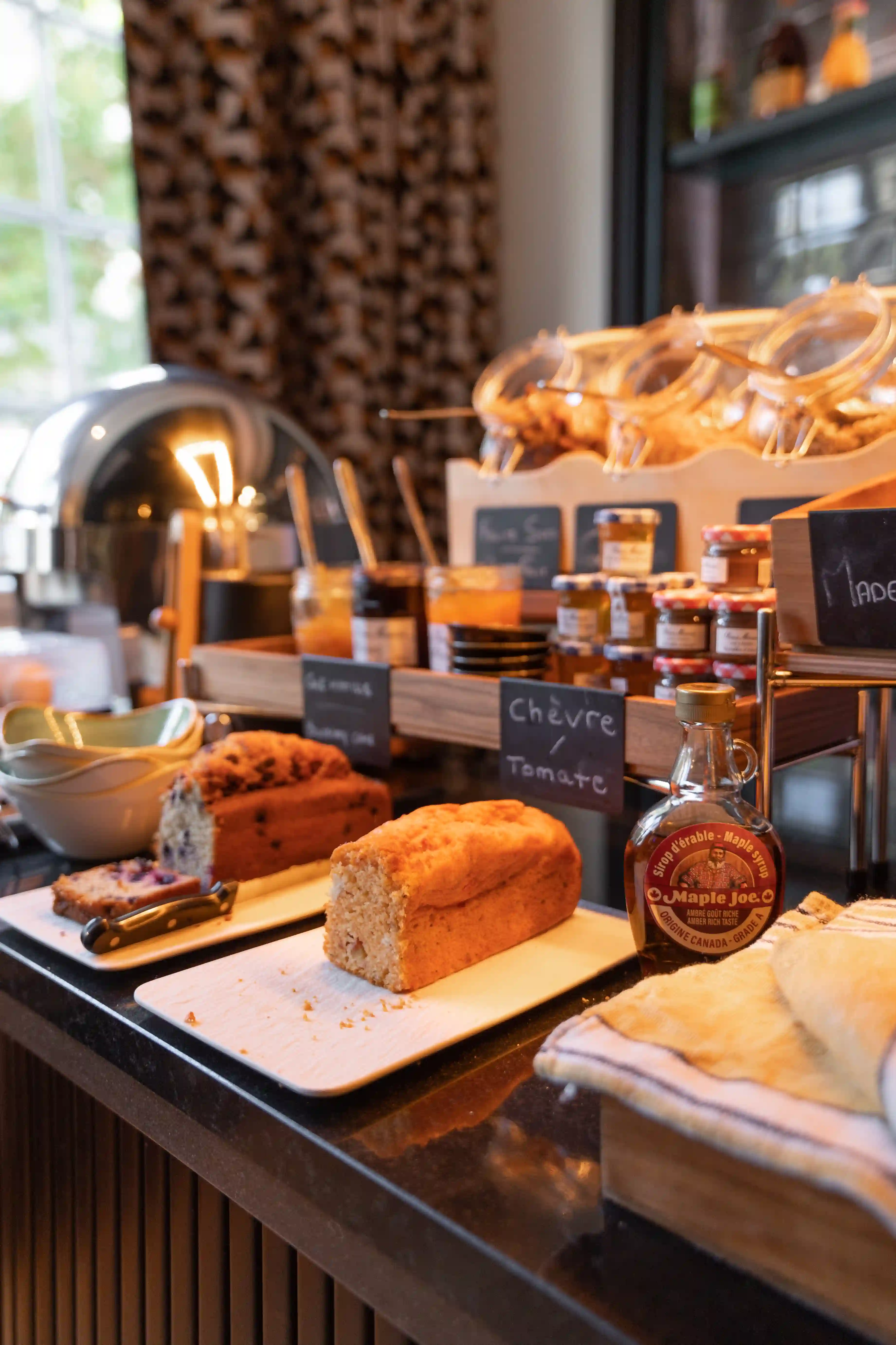 Plateau avec deux pains tranchés sur une table de buffet, un pot de sirop d'érable Maple Joe à côté, et des bocaux de confitures et miels en arrière-plan avec des étiquettes sur un présentoir en bois.