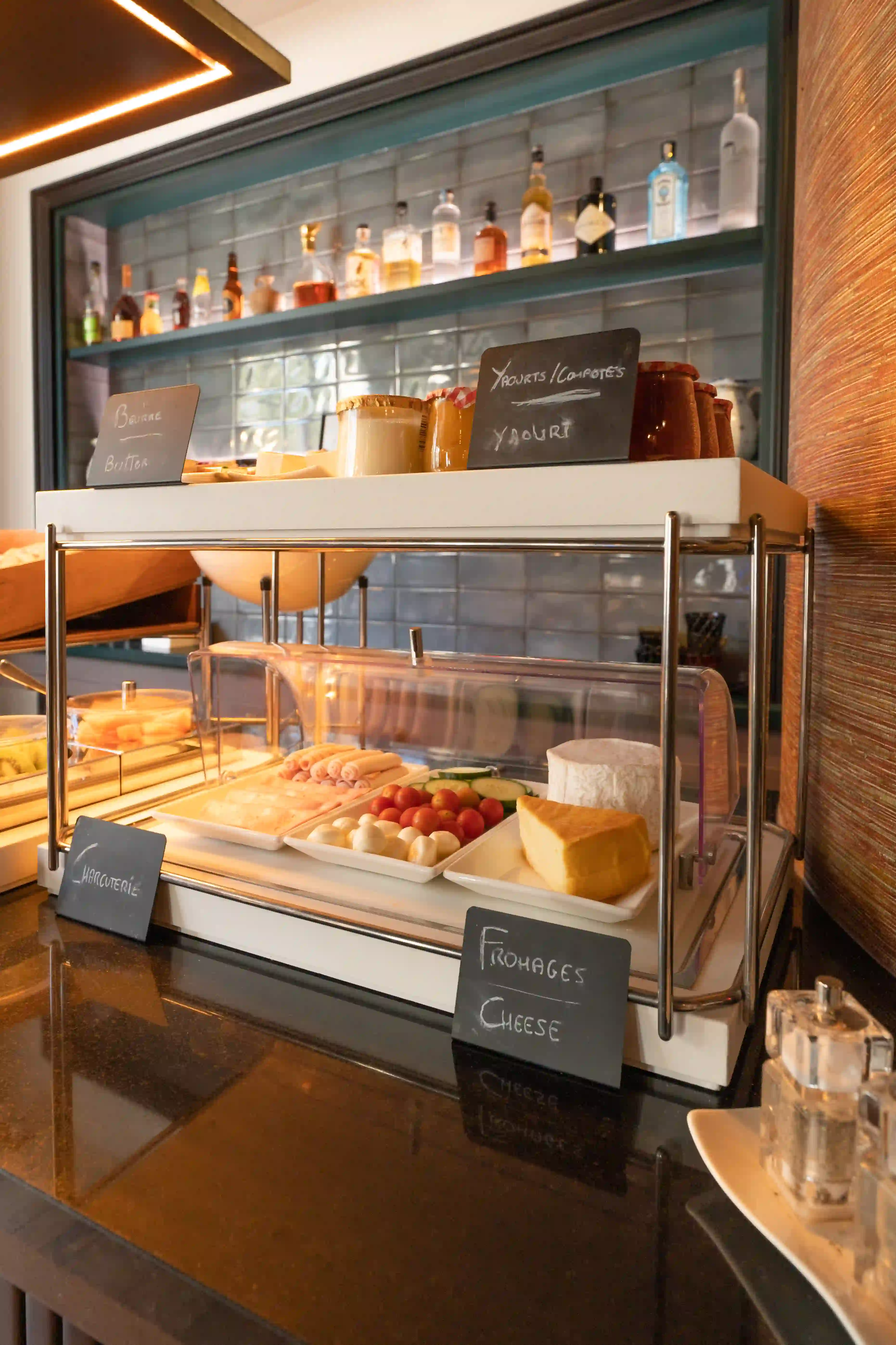 Vitrine réfrigérée affichant un assortiment de fromages, charcuterie, tomates cerises et légumes, avec des étiquettes indiquant beurre, yaourts, compotes, charcuterie et fromages, dans un cadre intérieur avec une étagère de bouteilles en arrière-plan.