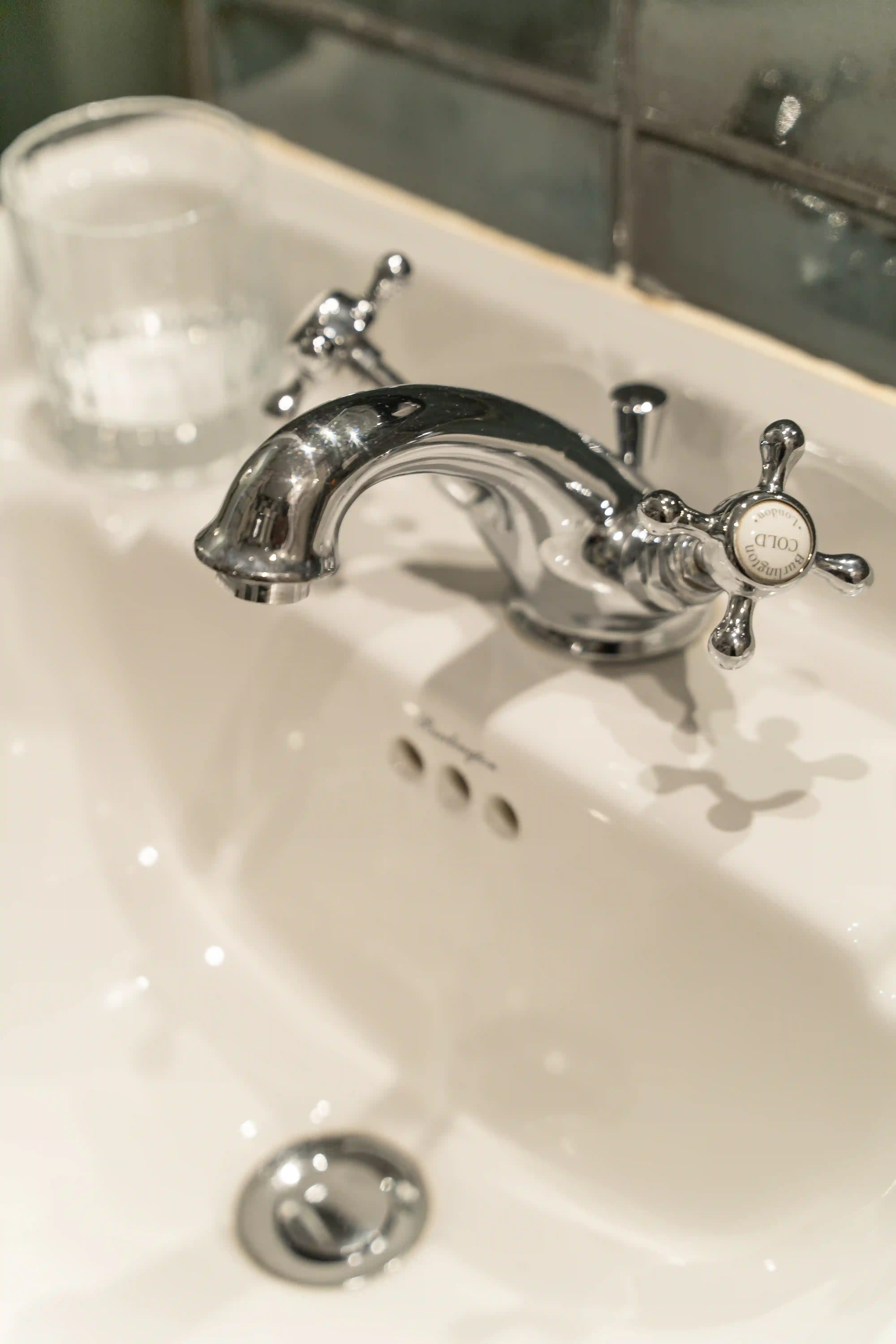 Torneira metálica cromada de comando duplo sobre um lavatório branco, com um manípulo marcado como frio e um copo transparente ao fundo, na borda do lavatório.