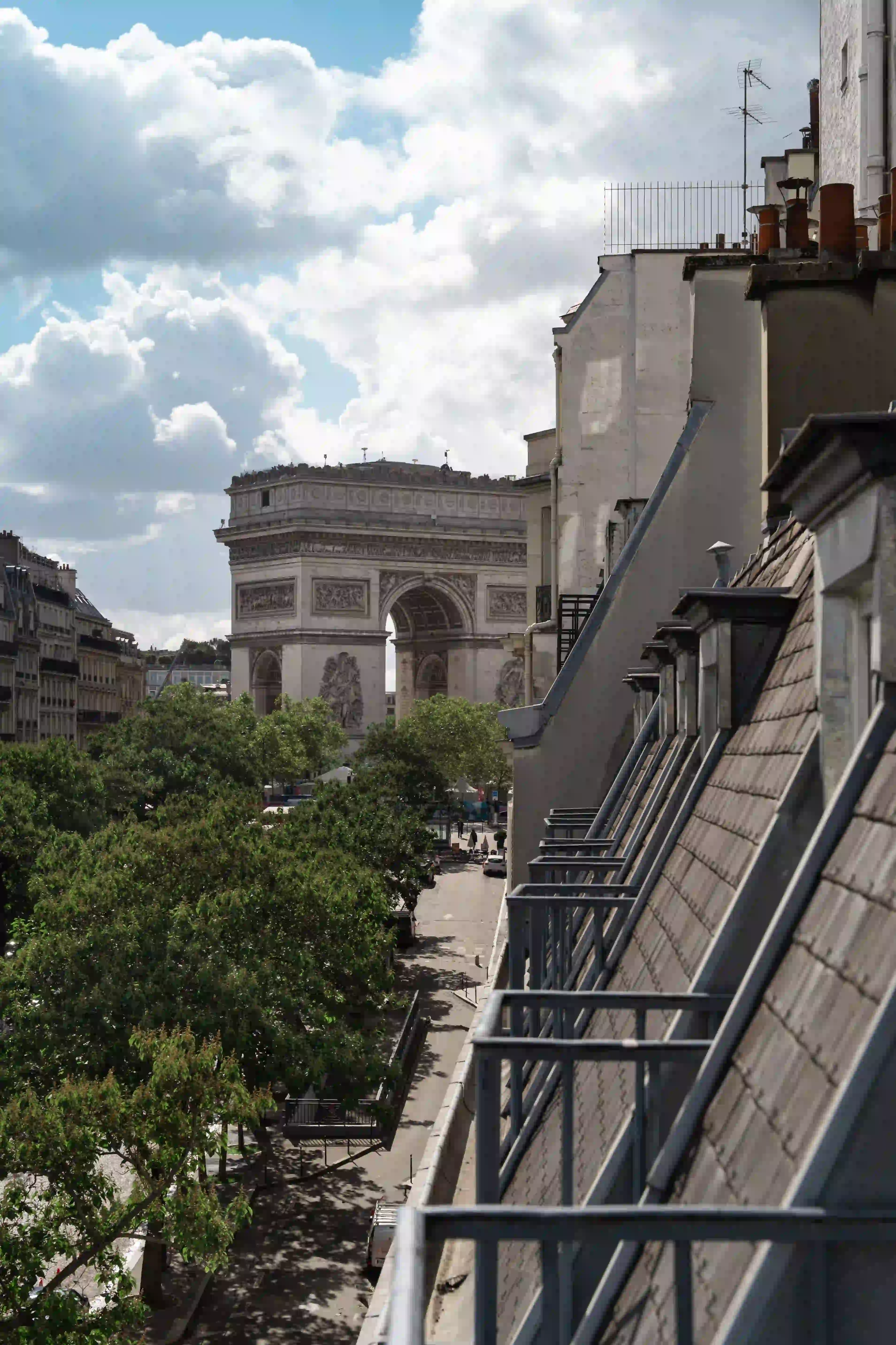 Vue sur l'Arc de Triomphe à Paris depuis un balcon, avec des toits en ardoise et des cheminées en premier plan, des arbres verts et une rue en dessous sous un ciel partiellement nuageux.