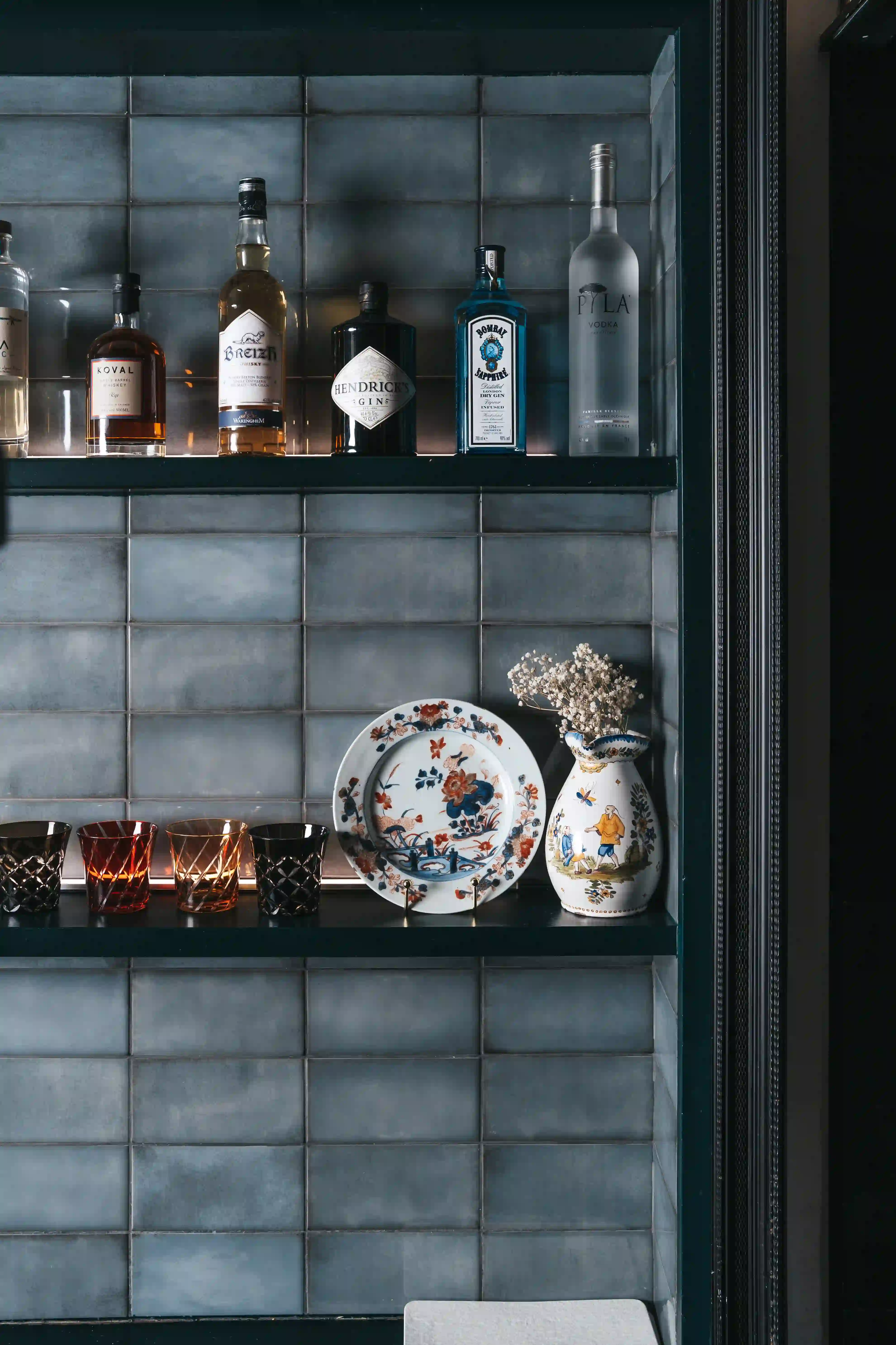 Prateleira de bar com várias garrafas de bebida na prateleira superior, quatro copos coloridos, um prato decorativo de faiança e um vaso ornamentado com padrões e flores secas na prateleira inferior, diante de uma parede de azulejos cinzentos.