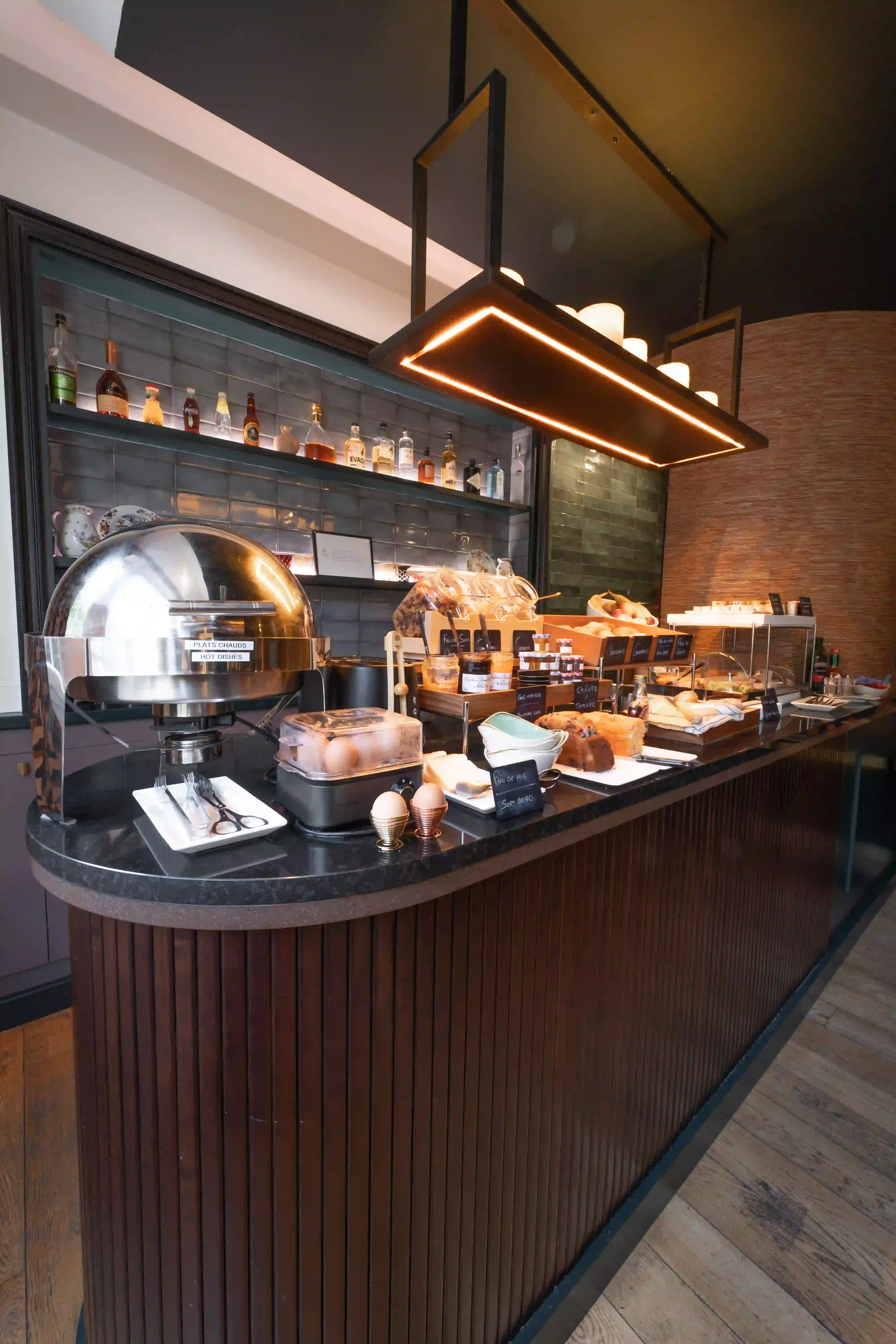 Buffet de pequeno-almoço com pratos quentes em aço, ovos cozidos, pães variados, compotas, pastas para barrar, tabuleiros de pastelaria e garrafas de bebidas alcoólicas numa prateleira ao fundo, num ambiente moderno.