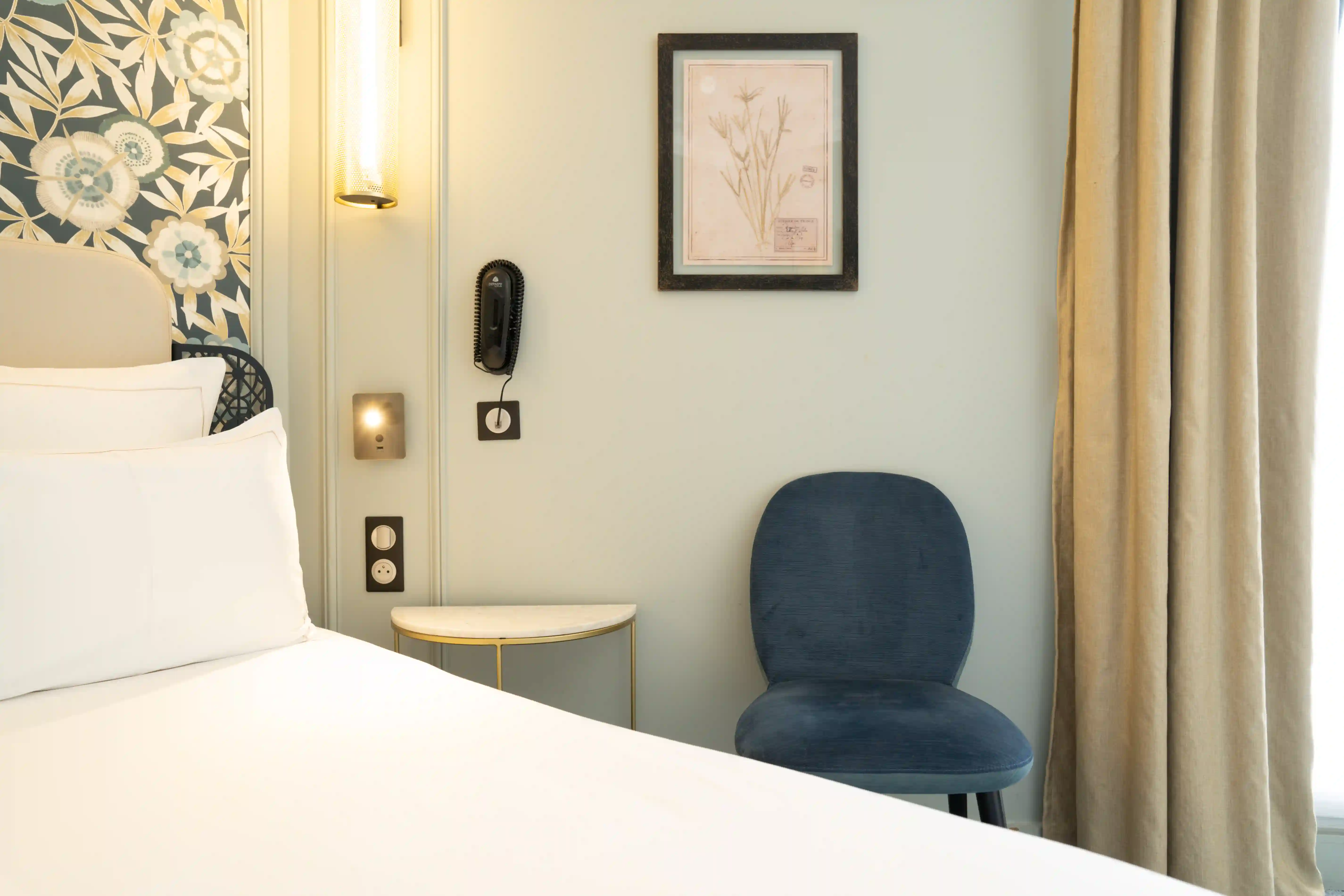 Canto de um quarto com uma cama branca, uma mesa de cabeceira redonda com tampo branco e pés dourados, uma cadeira azul e um quadro botânico emoldurado pendurado na parede clara, um candeeiro de parede iluminado e um telefone preto na parede, cortinas bege à direita.